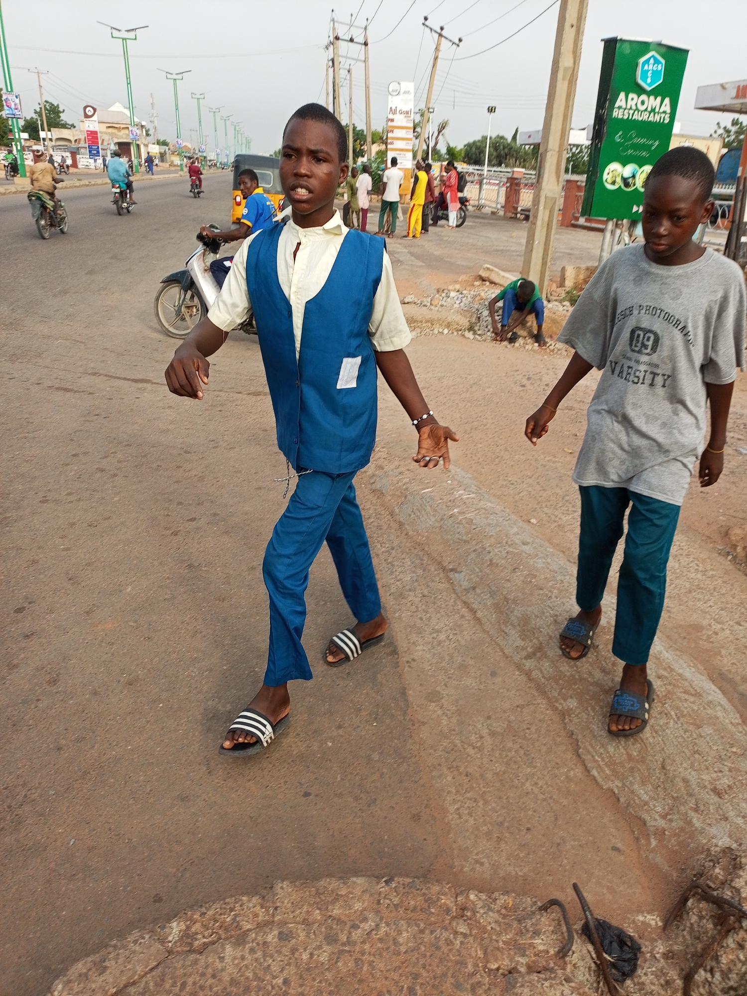 In the city of Gombe, Nigeria, two young boys are walking on a paved road. The boy in the foreground is wearing a light yellow shirt, a blue vest, and blue pants. He has dark skin and short hair, and is looking towards the viewer. He is wearing black and white striped sandals. The boy to his right is wearing a grey t-shirt with "09 VARSITY" printed on it and teal pants. He is also wearing sandals. In the background, other people and vehicles, including motorcycles and a tricycle, are visible on the road. Traffic lights and streetlights line the street, and signs for "AROMA RESTAURANTS" are also visible. The environment appears to be an urban street with some greenery and buildings in the distance.