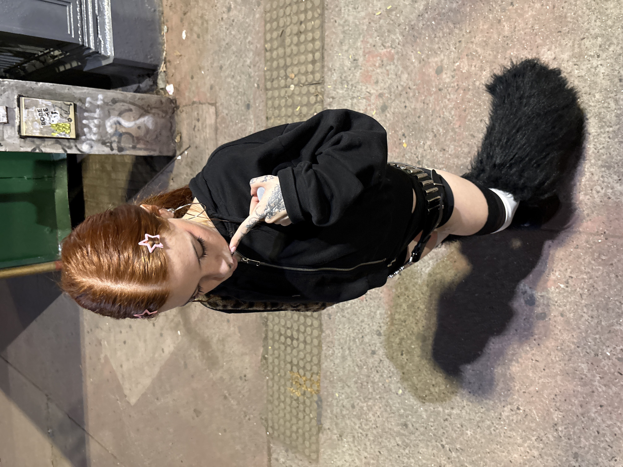 A high-angle shot depicts a red-haired person with pink star-shaped hair clips, wearing a black hoodie, black shorts with a studded belt, black and white striped socks, and shoes, standing on a paved surface. The person is touching their face with a tattooed hand. A black furry tail is attached to their clothing. In the background, there is a wall with graffiti and a green object. The location is likely an urban area, possibly in Buenos Aires, Argentina, but there is nothing in the image to confirm that.