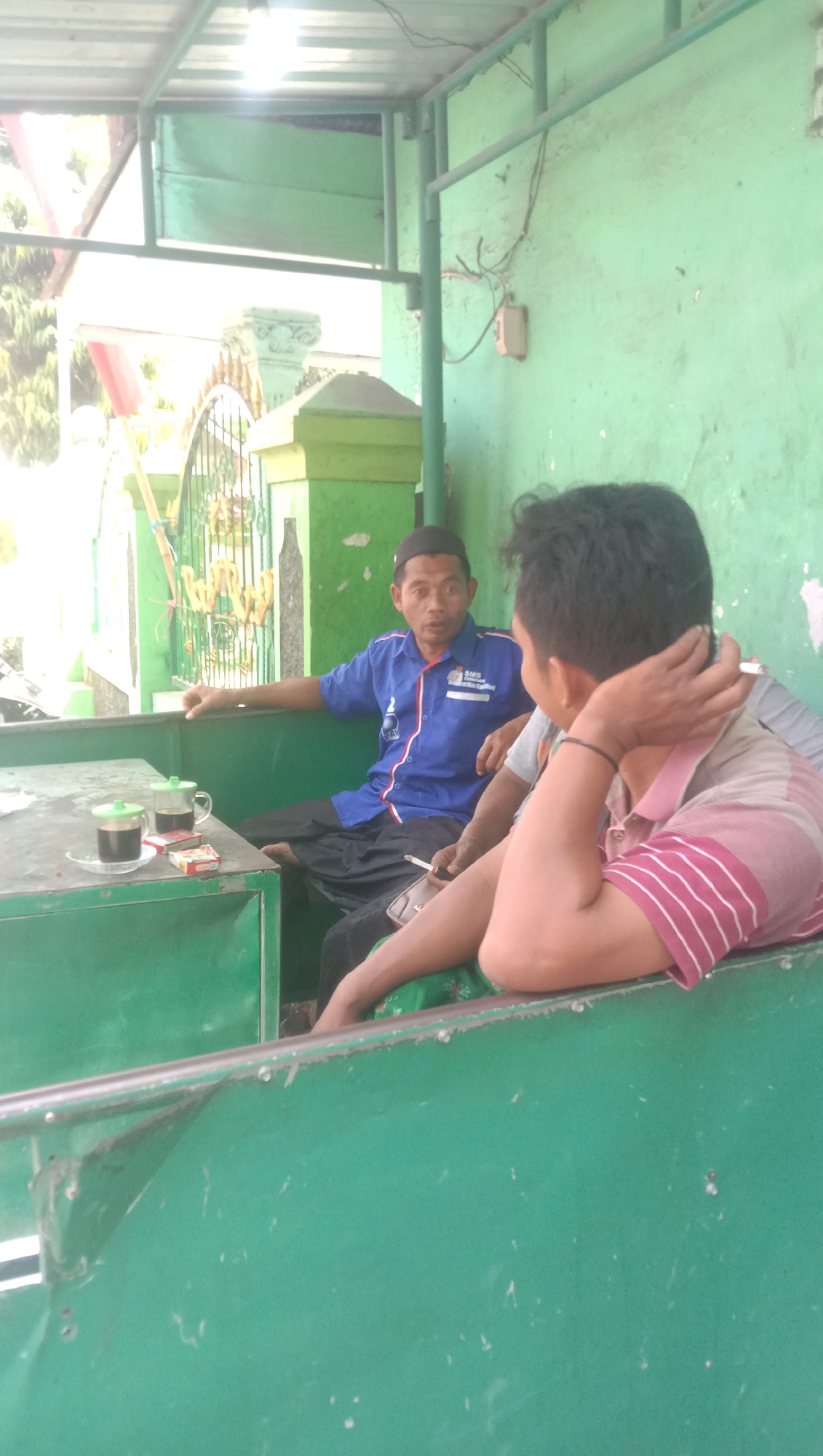 In Karanganyar, Indonesia, two men are seated at a green metal table under a corrugated metal roof. The man on the left wears a blue shirt with red and white trim and a black skullcap. He holds a cigarette in his right hand and gestures with his left. He is barefoot and wears black pants. On the table in front of him are two cups of coffee, a small pack of cigarettes, and a box of matches. The man on the right, partially visible, wears a striped shirt and has his arm resting on his forehead. He is also smoking a cigarette. The background is a green wall, and a decorative metal gate and gatepost are visible through an opening to the left. The scene appears to be an outdoor seating area, possibly a cafe or a roadside rest stop.