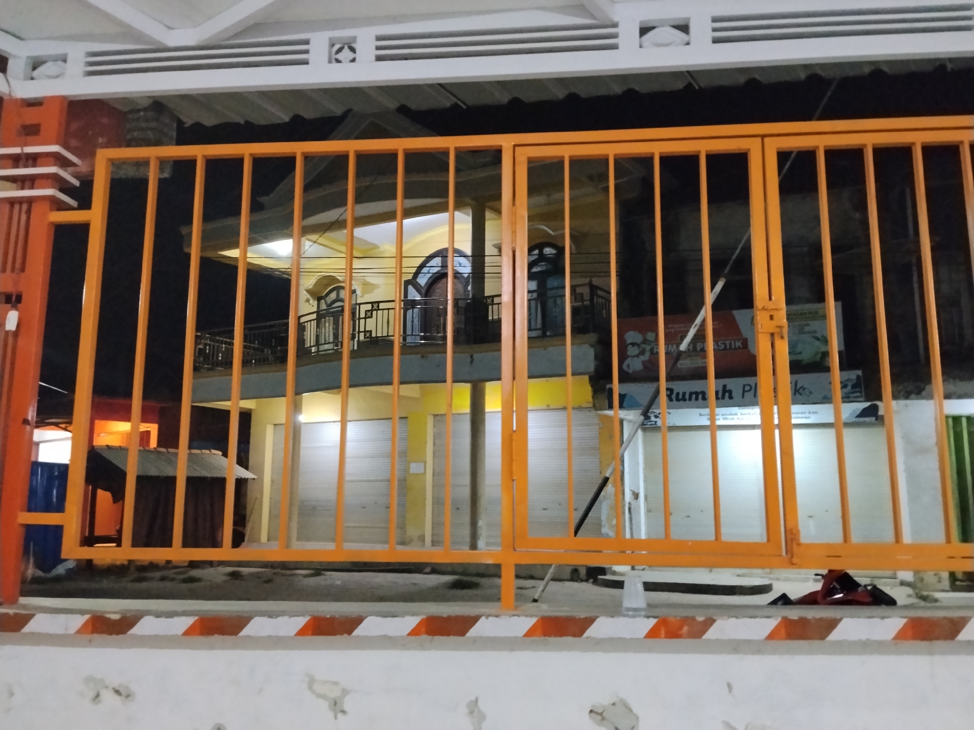 The image captures a brightly lit building with an orange metal gate in the foreground. The building appears to be a multi-story structure, with the lower level featuring shuttered windows or doors. Above the shutters, a yellow wall is visible, and on the second level, there is a balcony with black metal railings. The building is illuminated by artificial lights, creating a warm glow. A small, dark-colored shed or structure is partially visible to the left of the gate. The overall scene suggests an establishment in a commercial or residential area, likely at night, given the lighting conditions.