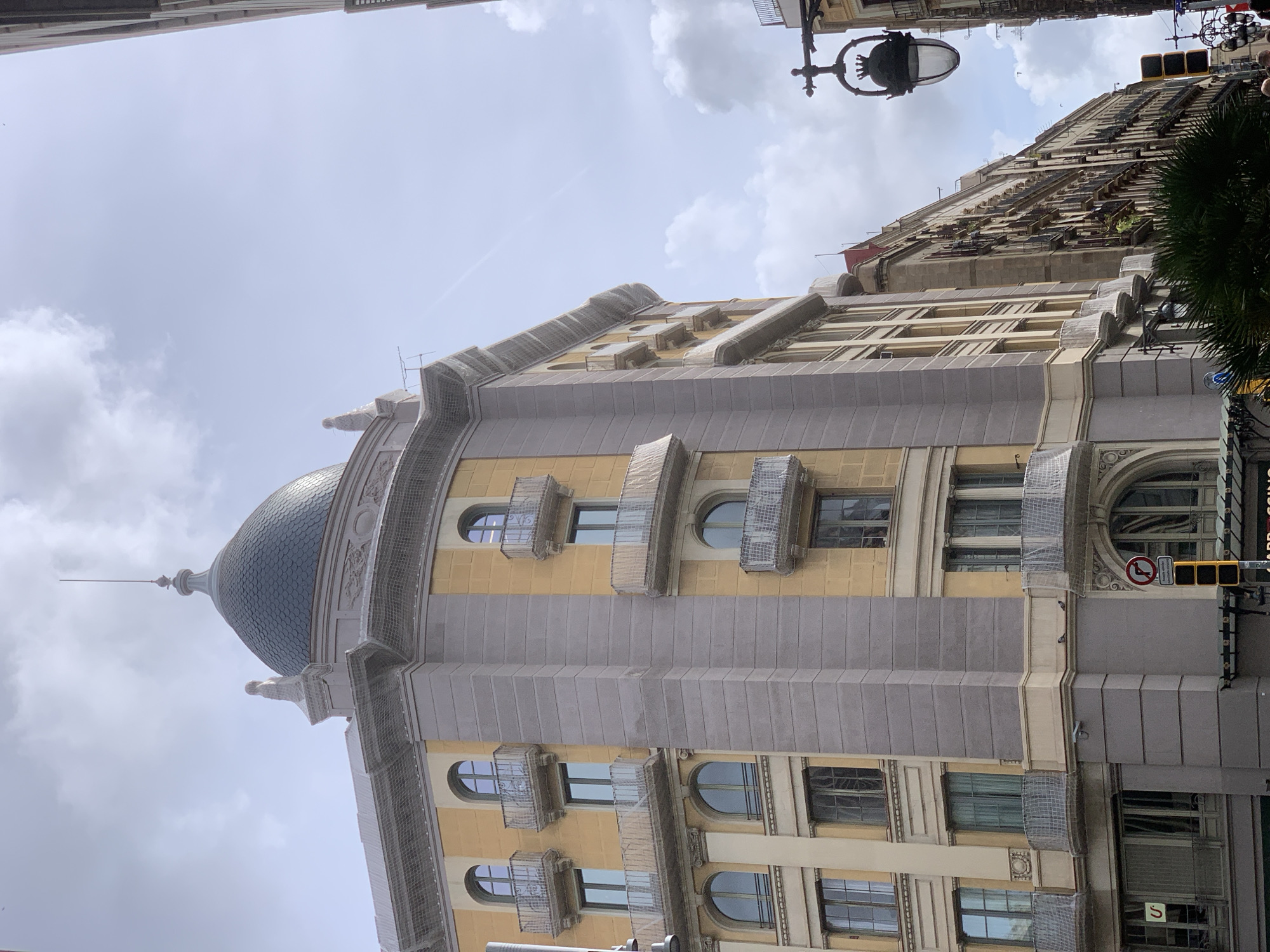 The image shows the facade of a building in Barcelona, Spain. The building is characterized by a dome on its left side and a series of balconies on the facade. The color scheme of the building consists of shades of yellow and grey. The architectural style appears to be in the Art Nouveau or Modernista style, typical for buildings designed by Antoni Gaudí. A traffic light is visible in the bottom right corner. Additionally, the facades are covered with netting.