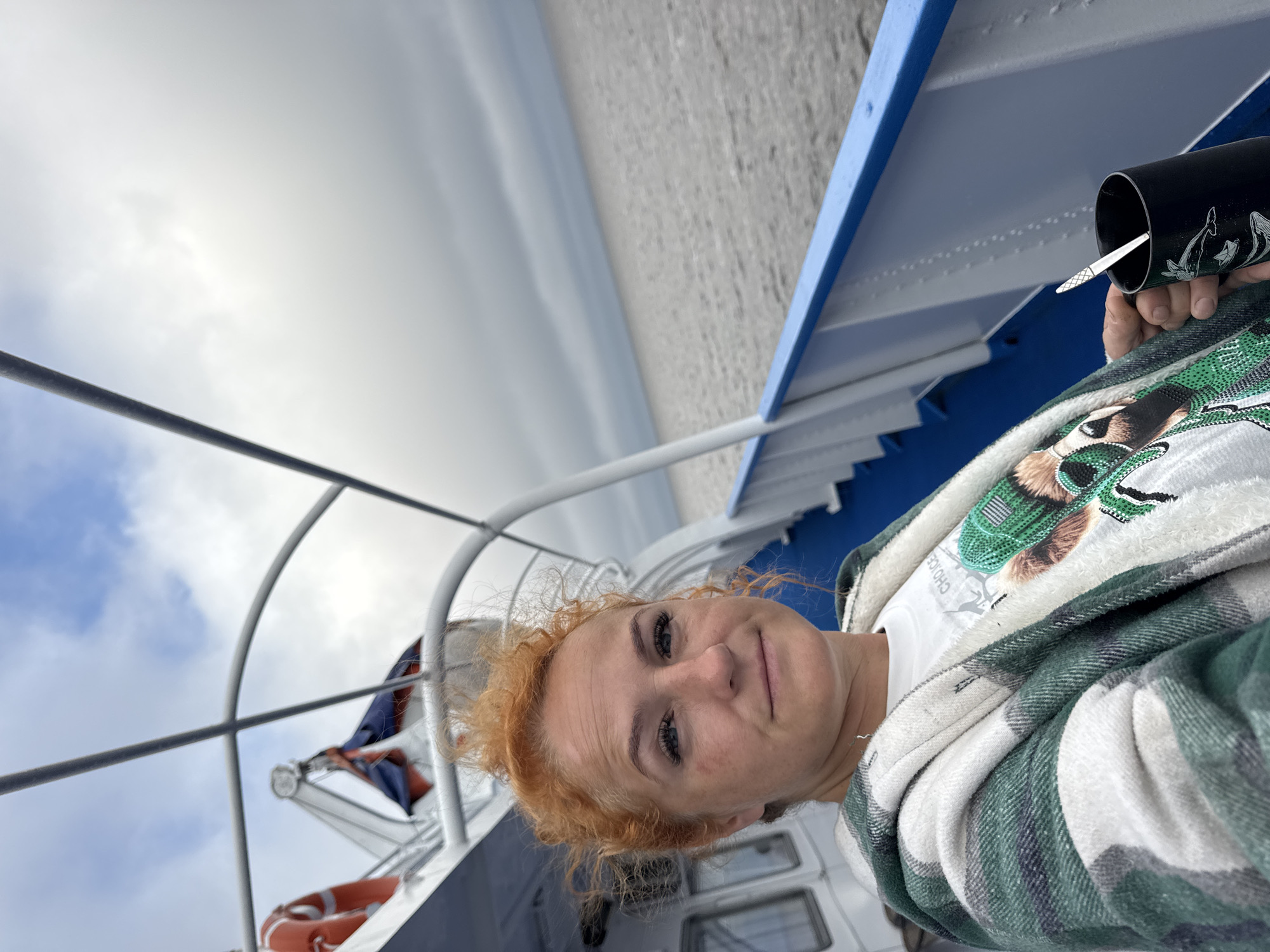 In Laishevo, Russia, a woman with reddish-orange hair, styled in a messy bun, is photographed from the chest up on the deck of a boat. She is wearing a white t-shirt with a graphic design and a green and white plaid fleece jacket. Her left hand is holding a black mug with a spoon in it, featuring a design of dolphins. The woman is looking towards the camera with a neutral expression, and her makeup is visible. The background shows the railing of the boat, the sky with clouds, and the water. Life preservers are visible on the deck, suggesting safety equipment is present. The overall atmosphere is casual and suggests a journey on the water.