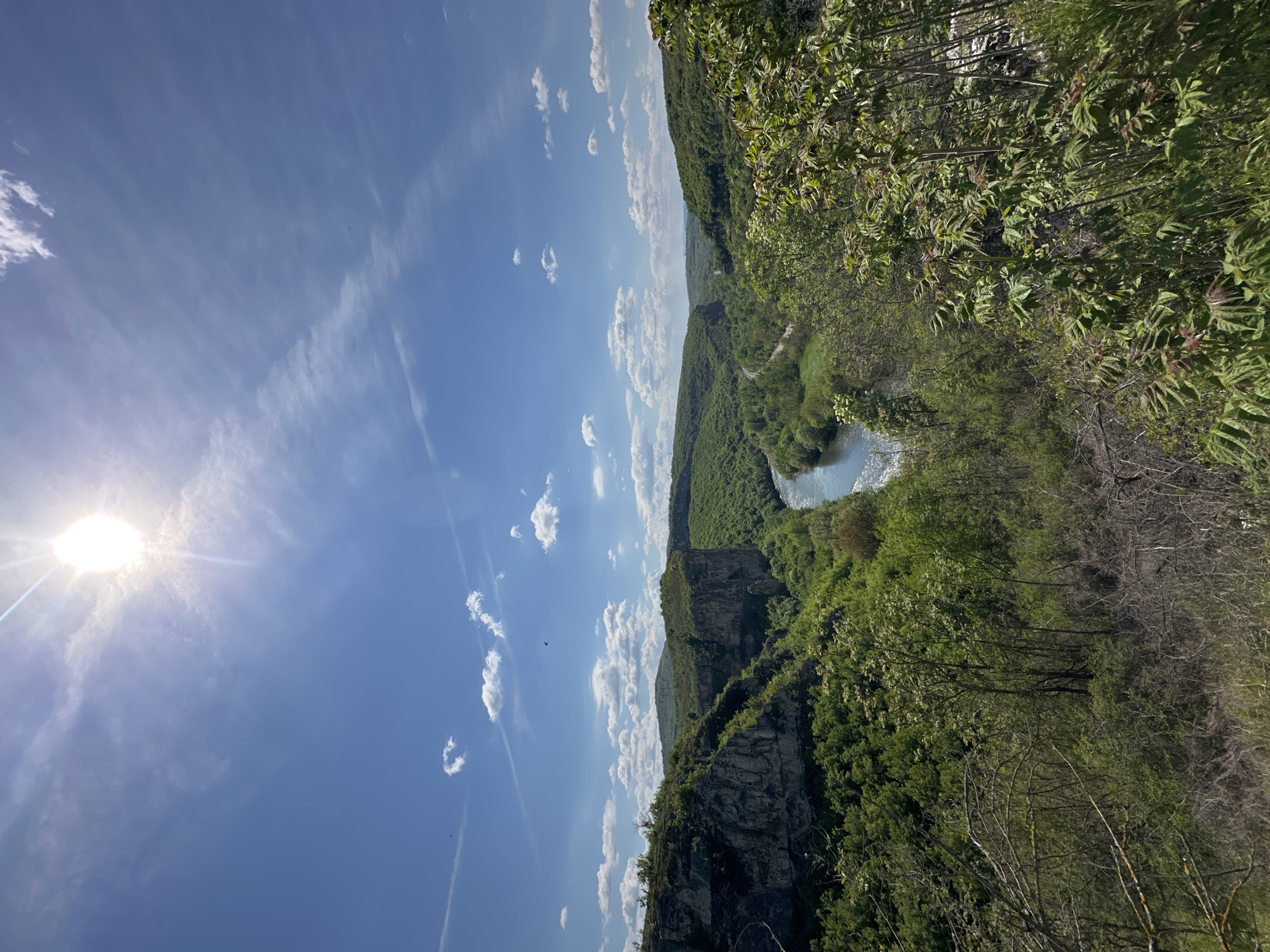 The image shows a landscape view of Karlukovo, Bulgaria, featuring a river winding through a deep gorge. The scene is dominated by the steep, rocky cliffs and lush green vegetation covering the surrounding hills. The sky is blue with scattered white clouds. The sun is positioned on the left side of the image, creating a bright glare.