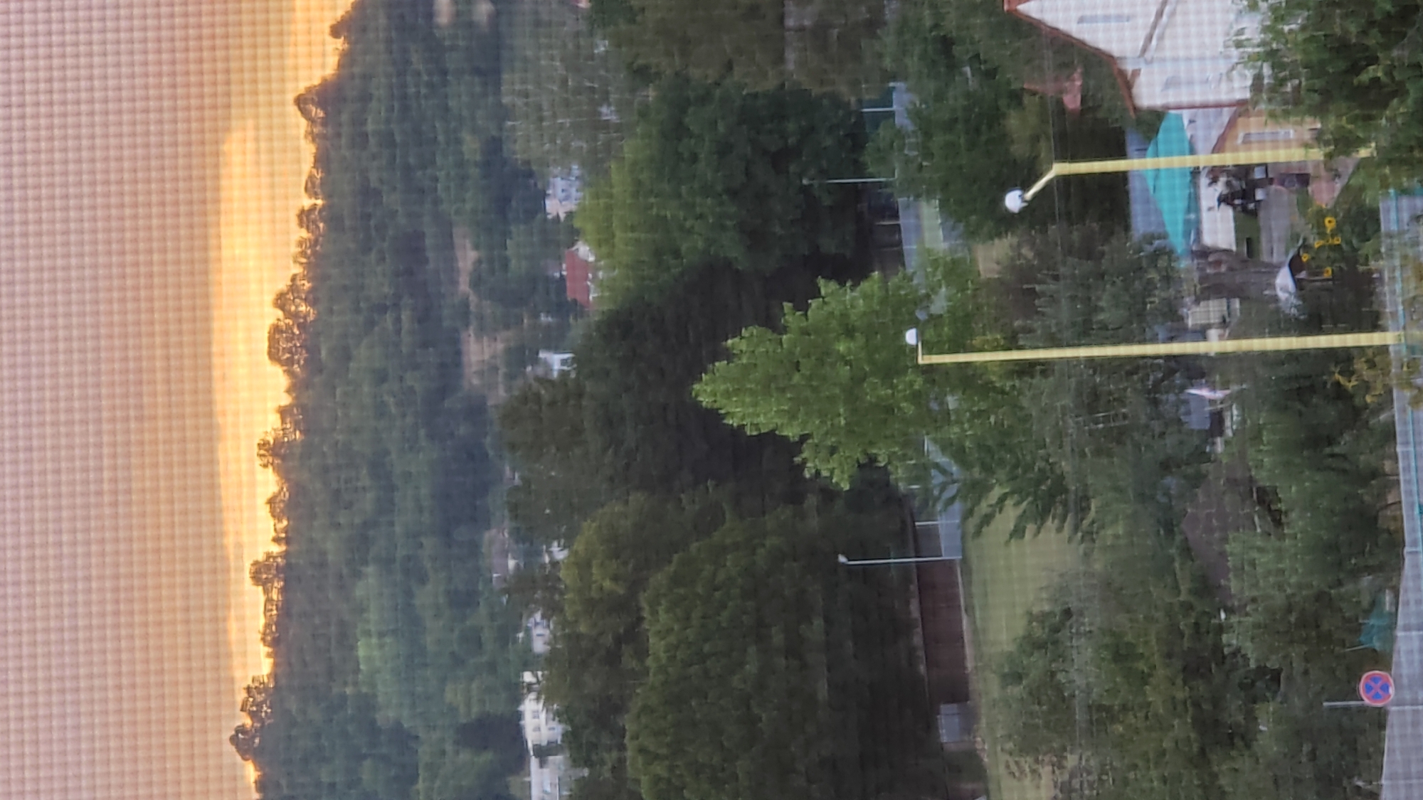 The image displays a hazy, panoramic view of a treed hillside with a soft, warm sky at dusk. The sky has a gradient of muted oranges and yellows, suggesting the sun is low on the horizon or has recently set. The hillside is densely covered with various shades of green trees, with a more pronounced, lighter green tree in the mid-ground that stands out from the rest.
In the foreground and mid-ground, there are glimpses of a residential area interspersed with more trees. Various houses with red and white roofs are visible, along with what appear to be streetlights. One prominent streetlight with a yellow pole and a white lamp head is positioned in the right mid-ground. Another similar streetlight is visible slightly to its left. A turquoise umbrella or canopy is also present in the mid-ground.
At the very bottom, a thin horizontal fence is visible, and to the left, a circular traffic sign with a red border and blue cross can be discerned. The overall impression is a tranquil, somewhat blurred outdoor scene, likely captured from an elevated position overlooking a suburban or semi-rural area. The location is identified as Prešov, Slovakia.
