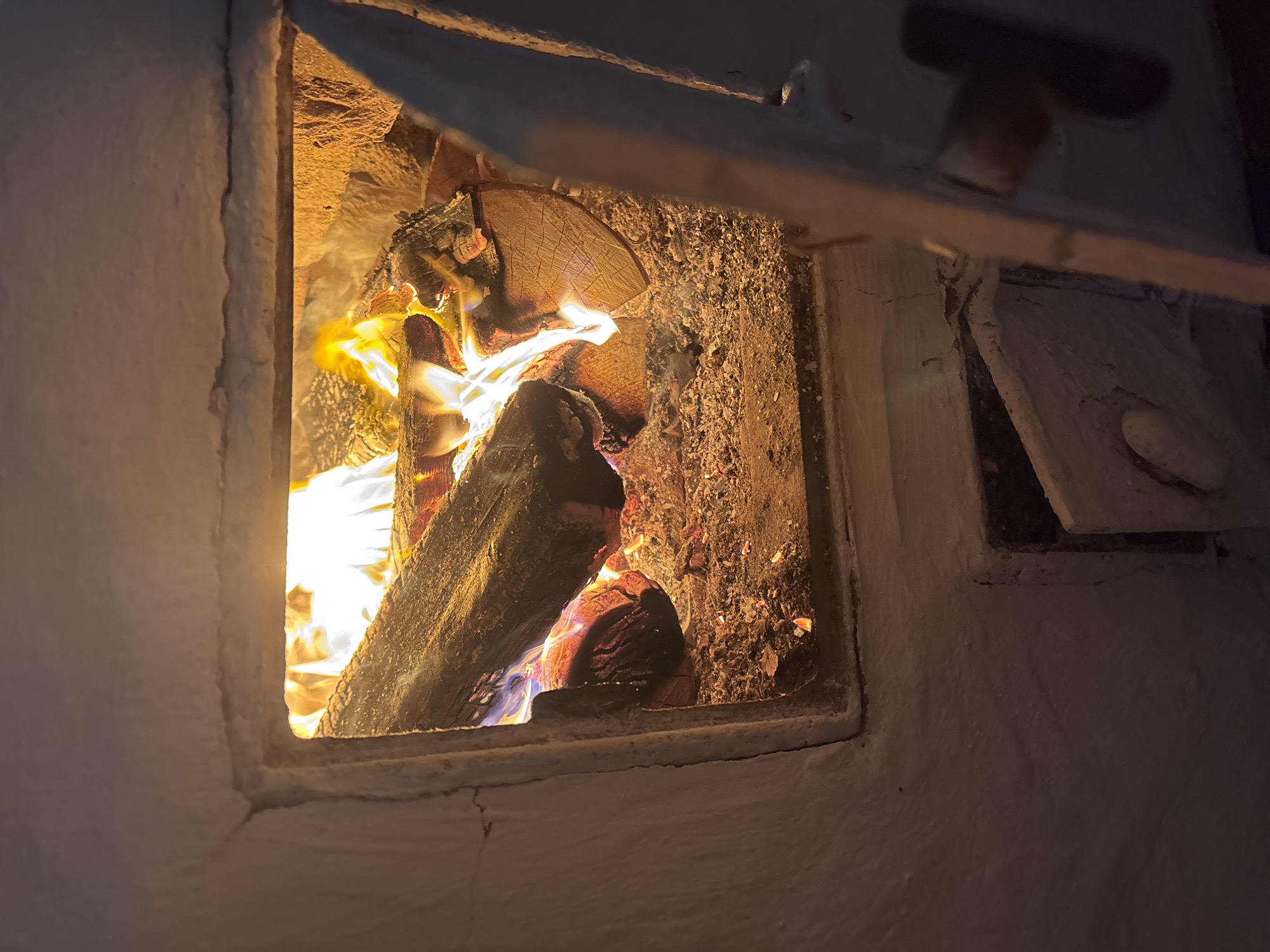 The image shows a close-up view of a burning fireplace or stove, likely made of white plaster or brick. Logs are actively burning inside, with visible flames, embers, and ash. The opening of the fireplace is rectangular. To the lower right, a small, open metal door, possibly for ash removal, is visible. The scene is dimly lit, with the primary light source being the fire itself. The image does not contain any information about the city of Курган, Russia, or any people.
