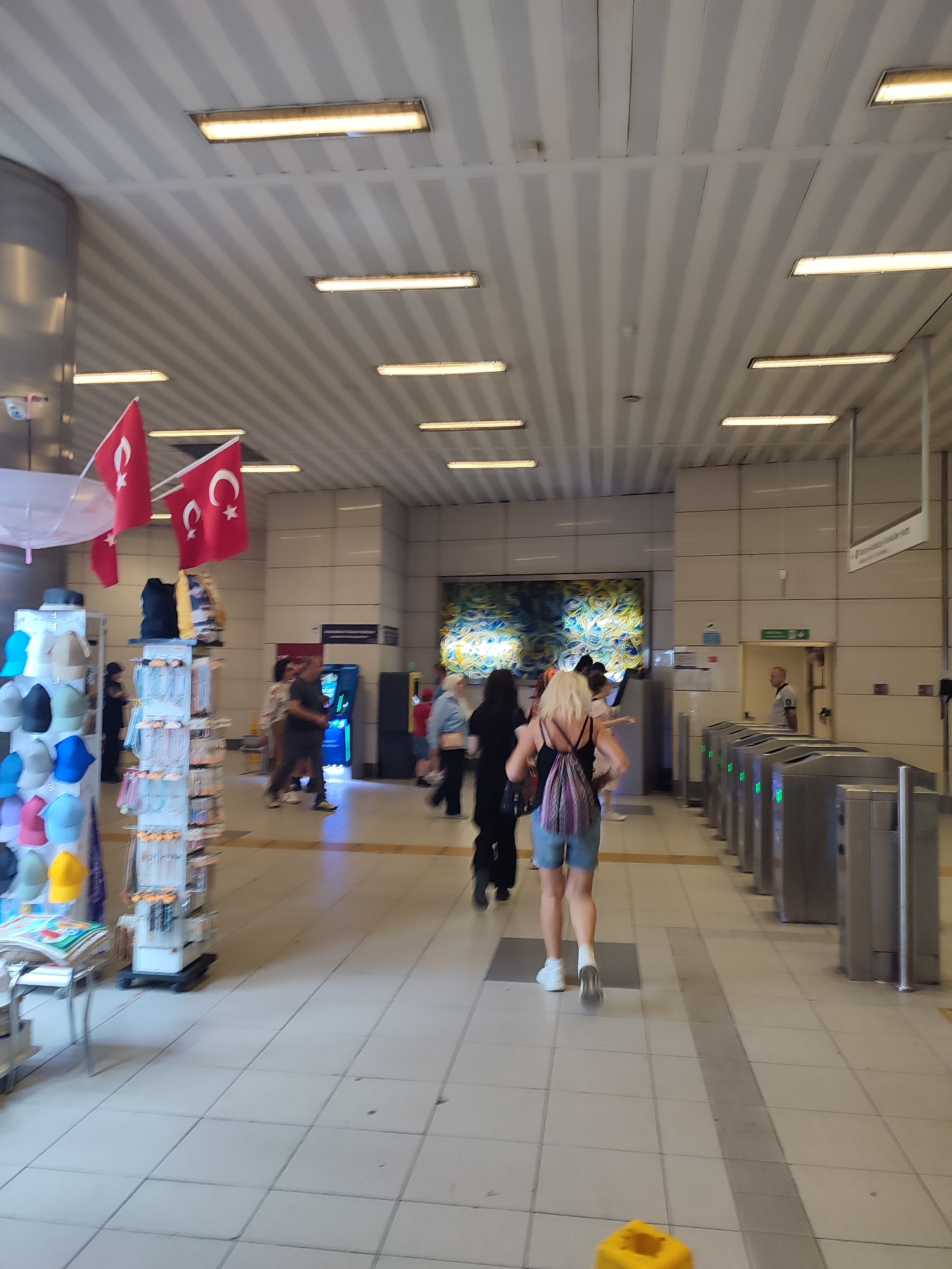 The image depicts an indoor public space, likely a metro station, in Beyoğlu, Turkey. The environment is characterized by a white, tiled floor and a ribbed, white ceiling with several fluorescent light fixtures.
In the foreground on the left, a display rack holds numerous colorful caps. Behind it, two Turkish flags are prominently displayed. Further back, several individuals are walking through the space. One woman, with blonde hair and wearing a black tank top and denim shorts, is walking away from the camera, carrying a patterned backpack. Another person in a black outfit is walking ahead of her.
To the right, a row of turnstiles with green illuminated indicators suggests an entrance or exit point. A man in a security uniform is visible standing near an open doorway in the background. A colorful mosaic artwork adorns the wall on the far side of the passage. The scene conveys a sense of everyday activity within an urban transit hub.