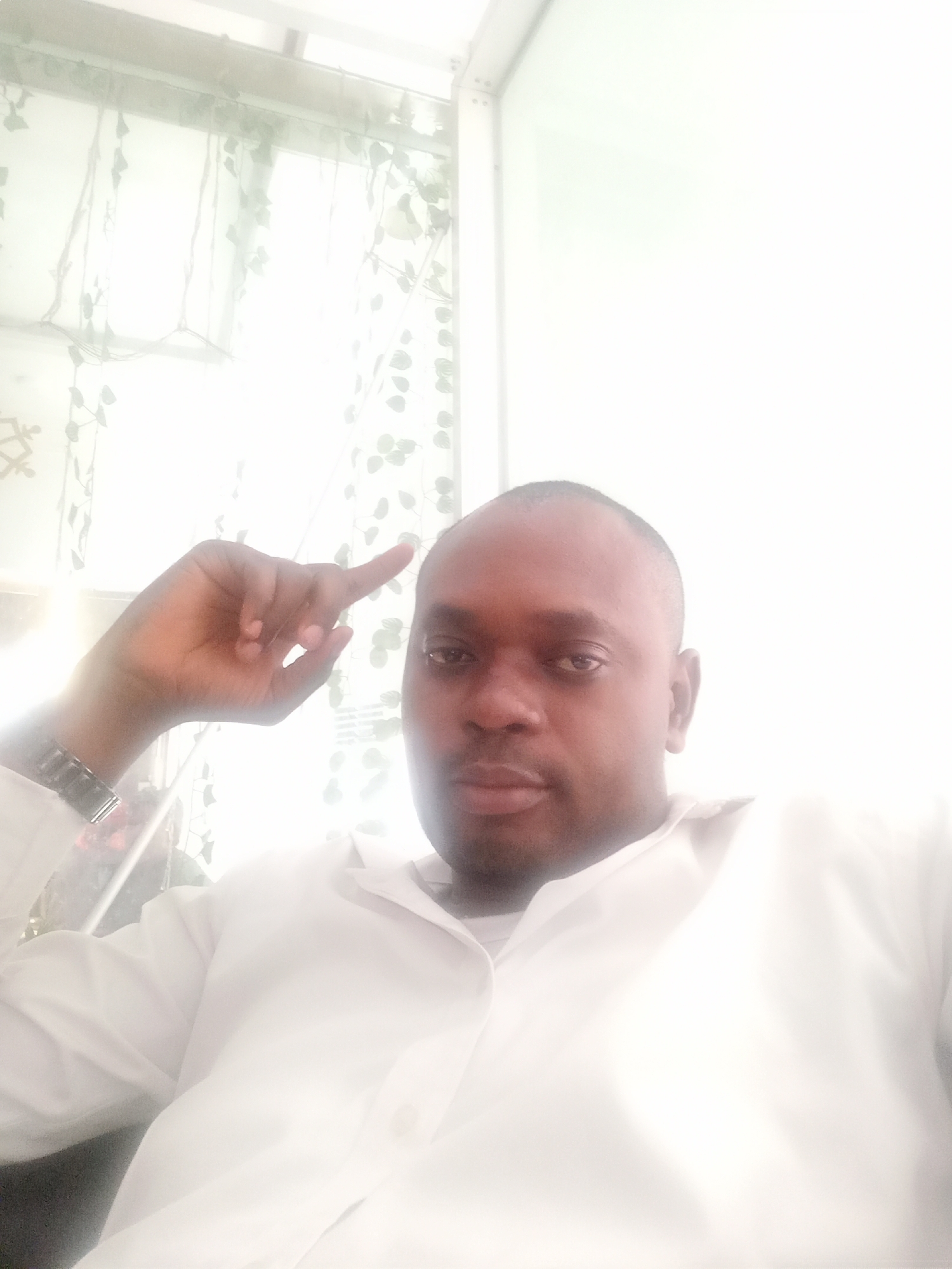 The image is a brightly lit, close-up selfie of a man taken indoors. The subject is a man with dark skin, short dark hair (with a visible receding hairline), and a faint beard or stubble. He is wearing a white, long-sleeved collared shirt. On his left wrist, he has a metallic watch. His left hand is raised in the frame, with his index finger pointing upwards or to the right. He looks directly into the camera with a neutral to serious expression.
The background is significantly overexposed due to bright light, likely from a window. Behind the man, a sheer curtain or decorative screen is visible, featuring a pattern of green leaves or vines. Hints of a window frame or structural elements are vaguely discernible in the upper and right portions of the background. The strong backlighting makes it challenging to identify further details of the room. The scene suggests an informal indoor setting during daytime due to the intense natural light. There is no visible text within the image. The location context is Gwarinpa, Nigeria.