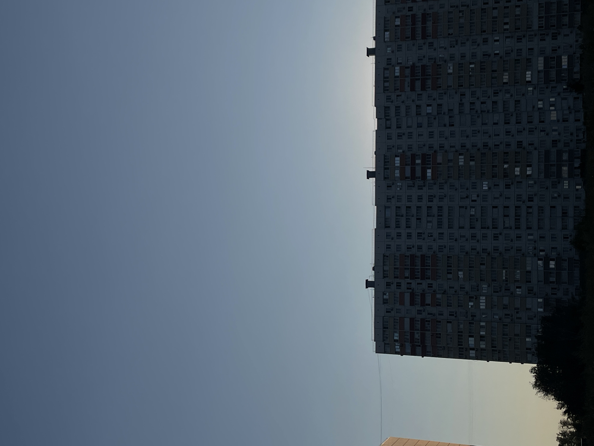 The image displays a multi-story apartment building silhouetted against a clear, pale blue sky. The building is a long, rectangular structure with numerous windows, many of which appear dark. Two chimneys are visible on the rooftop. To the left of the main building, a portion of another, lighter-colored building is partially visible. The lower portion of the image shows a dark silhouette of trees and foliage. The scene appears to be an outdoor urban environment, likely taken at dusk or dawn due to the lighting. The specific location in Rostov-on-Don, Russia, is not visually identifiable from the provided image alone.