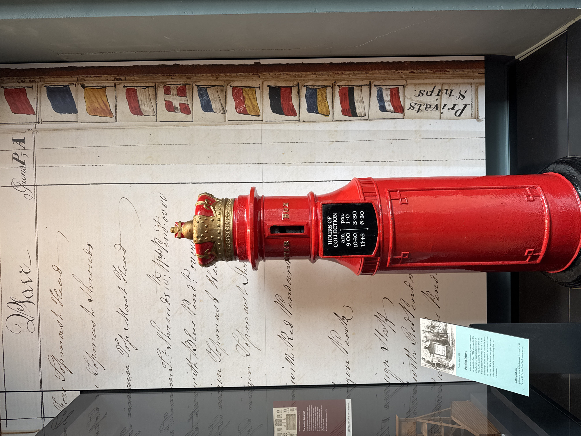 The image shows a red pillar box with a crown on top, positioned in front of a large display featuring handwritten text and small illustrations of flags. The pillar box has a plaque that reads "HOURS OF COLLECTION" with a schedule below it. To the left of the pillar box, there is an information panel with text and an illustration of an early pillar box. In the background, a large, weathered document with calligraphic handwriting and a series of small, colorful flags arranged vertically is visible. The overall setting appears to be a museum exhibit. The specific location in the City of Edinburgh, United Kingdom is the National Museum of Scotland.