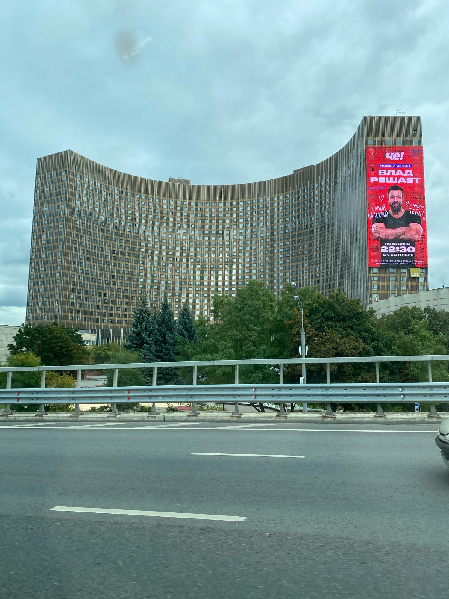 This image captures a view of the Cosmos Hotel in Moscow, Russia. The large, curved building dominates the frame, with its numerous windows characteristic of Soviet-era architecture. A prominent billboard advertising a television show, featuring a man and the text "Влад решает" (Vlad Decides), is displayed on the side of the building. The billboard also includes details about the show's timing and channel. In the foreground, a highway with white lane markings is visible, with a metal guardrail separating the road from the embankment. Trees and greenery are present between the hotel and the highway, suggesting an urban park or landscaped area. The sky is overcast, indicating a cloudy day. The image appears to be taken from a moving vehicle.