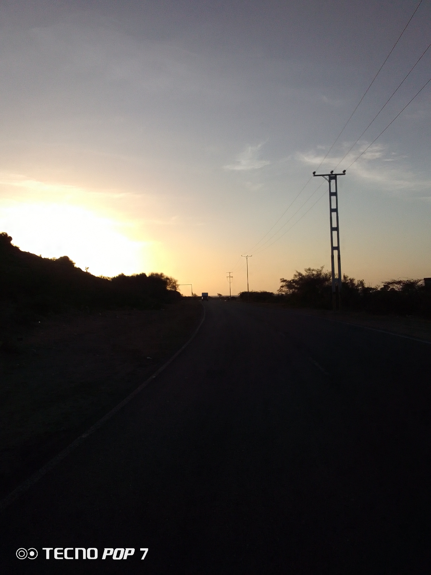 The image shows a road leading into the distance at dusk. The sky is bright with the setting sun on the left, fading into a pale blue on the right. There is vegetation on both sides of the road, silhouetted against the light. Utility poles line the right side of the road, with wires extending across the frame. In the distance, a vehicle can be seen on the road. The road surface is dark, with a faded line marking the edge. In the lower left corner, "Tecno POP 7" is visible. It is not possible to determine the specific location as the images are limited.
