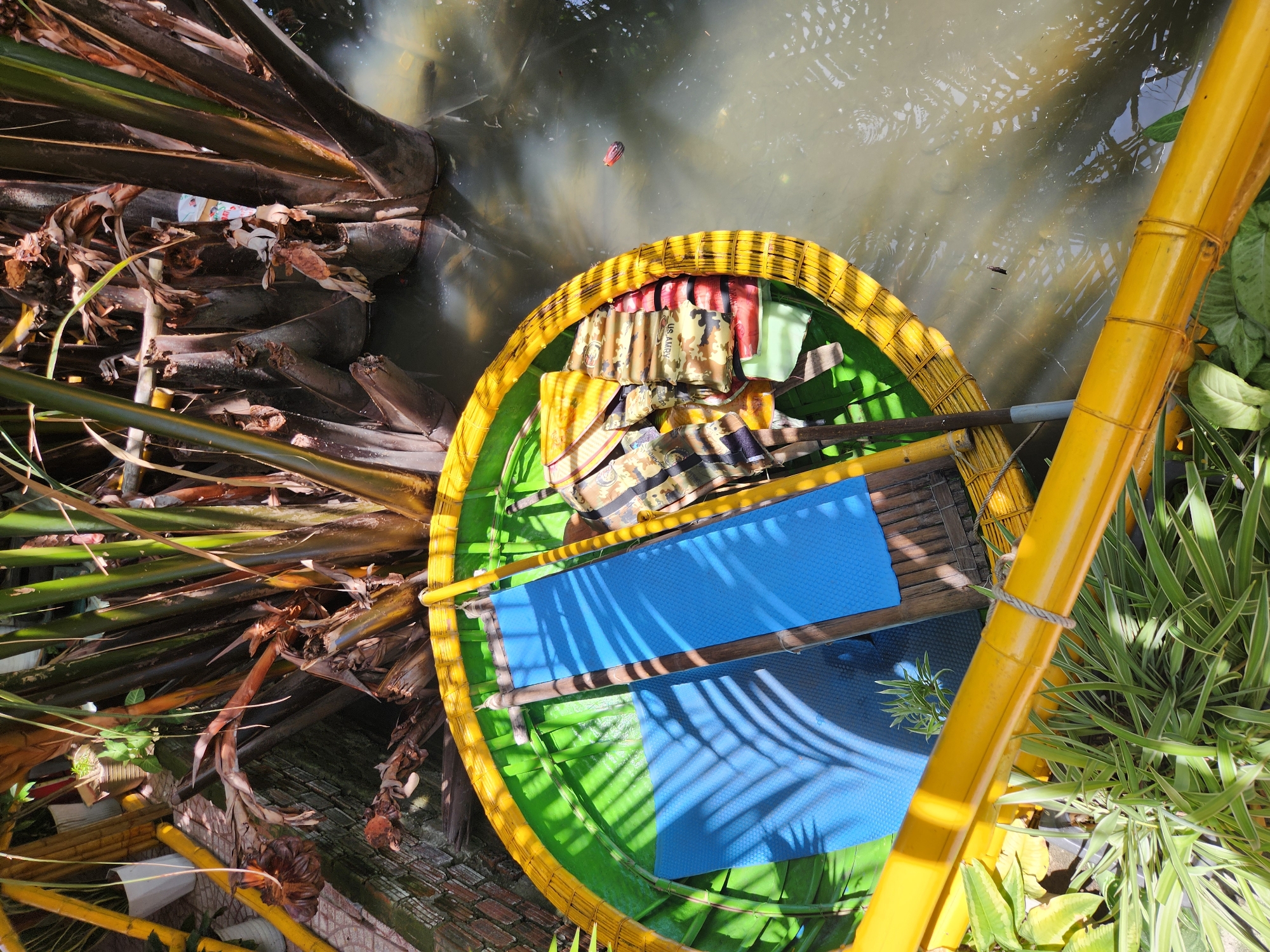 This is a high-angle, close-up, outdoor, natural light photograph taken in Da Nang, Vietnam, depicting a traditional Vietnamese basket boat floating in murky water amongst dense vegetation. The boat is circular with a bright yellow rim and a green interior. Inside the boat, two blue mats and several stacked life vests with camouflage patterns and the "US ARMY" marking are visible. Two wooden poles, likely oars, are also present. The boat is positioned in a shallow body of water, surrounded by the thick, dark trunks and leafy fronds of palm trees, their shadows creating dappled patterns on the boat's interior. Yellow bamboo poles run along the foreground and partially frame the scene, intertwined with lush green plants. The water's surface is slightly rippled and reflects the surrounding foliage. A small, dark object floats in the water to the right of the boat.
