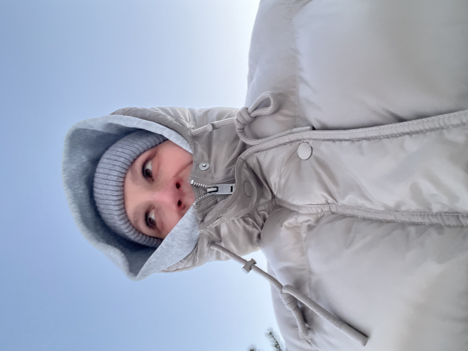 An individual is depicted in a low-angle, upward-looking shot against a clear, light blue sky in Putilkovo, Russia. The subject is wearing a light gray ribbed knit beanie under a light gray hooded sweatshirt, with the hood pulled up. Over these layers, a glossy, light beige/taupe puffy winter jacket is worn, zipped up to the lower chin. The jacket features a high collar, visible snap buttons, and drawstrings with toggles on either side. Only the upper portion of the subject's face is visible, including the eyes, forehead, and bridge of the nose, with the eyes directed upwards and to the right. The visible skin appears slightly flushed. A blurred, light-colored, possibly snow-dusted natural element is faintly discernible on the far left edge of the frame.