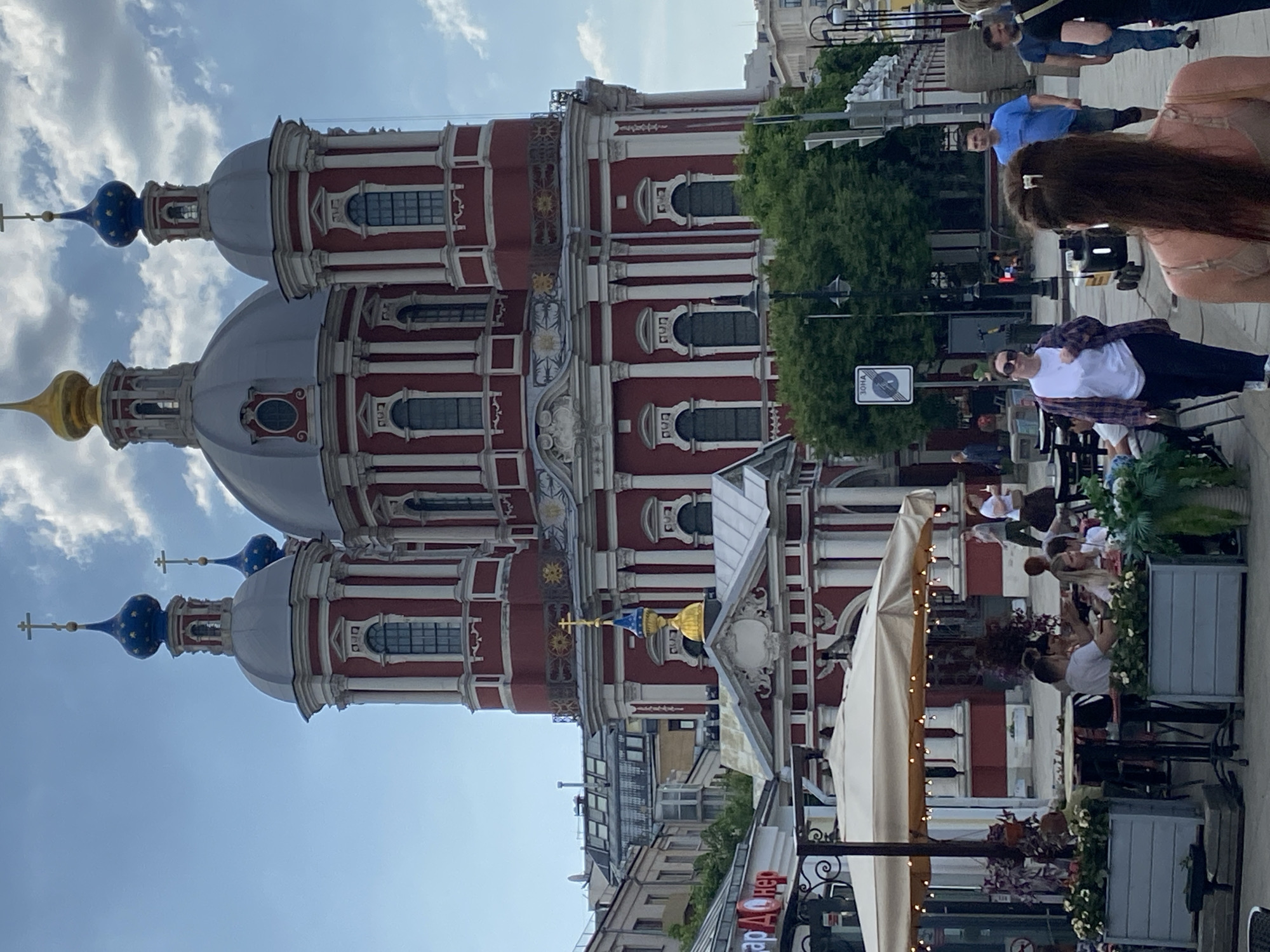 The image shows the Vysoko-Petrovsky Monastery in Moscow, Russia. The monastery's red and white buildings with distinct domes are visible in the background. Several people are present, some appearing to be walking along a pedestrian area, while others are seated at outdoor tables, likely part of a cafe or restaurant. There are awnings and planters near the seating area. The sky is partly cloudy, suggesting a sunny day.
