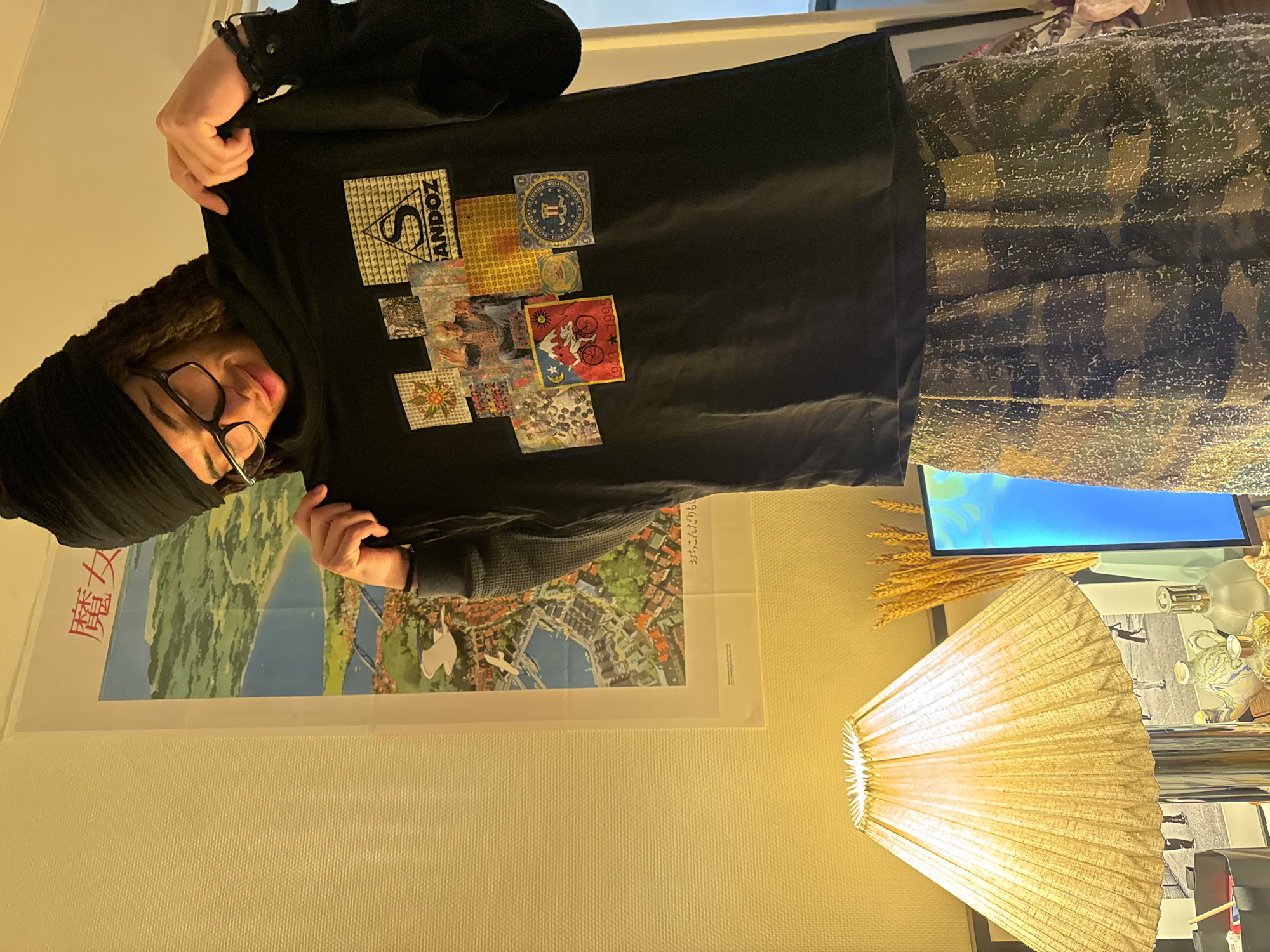 An individual, presumed male, with dreadlocks and wearing a black beanie and black-rimmed glasses, stands indoors holding up a black t-shirt. He is wearing a black long-sleeved shirt and camouflage-patterned trousers with a textured, possibly glittery finish. The black t-shirt displays a collage of colorful square and rectangular graphics, which include a triangular logo with "SANDOZ" text, a "DEPARTMENT OF JUSTICE FEDERAL BUREAU OF INVESTIGATION" seal, a psychedelic pattern, and a graphic featuring a figure on a mountain bike with "1943 * 1995". The individual looks down at the t-shirt.
Behind the individual, a large poster depicting a vibrant, idyllic coastal European-style town with hills, a bay, red-roofed buildings, and flying white birds is affixed to a textured, light-colored wall. Japanese text, including "魔女" (Witch) at the top, is visible on the poster, identifying it as artwork from Studio Ghibli's *Kiki's Delivery Service*. To the left, a table lamp with a pleated, light-colored fabric shade and a metallic base illuminates a surface below. This surface holds various objects, including a small black container, a ceramic teapot, a clear glass bottle, a bundle of dried wheat stalks in a vase, and a portion of a screen displaying blue light. A framed black and white image is partially visible behind the lamp. The scene takes place in an indoor setting, presumed to be in the city of Paris, France.