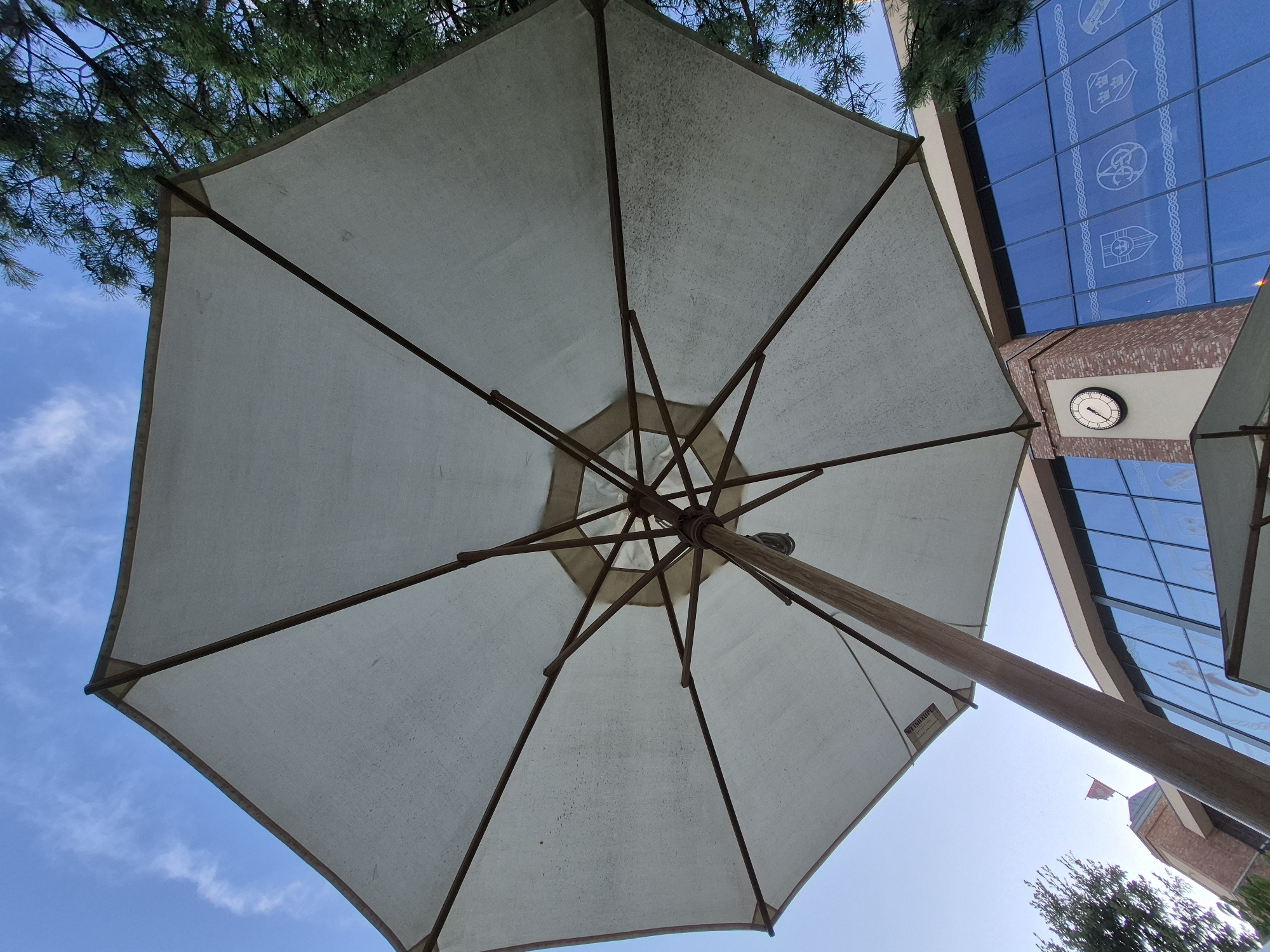 An under-view of a large, octagonal, light-colored outdoor umbrella with a wooden pole and ribs is captured against a bright blue sky with scattered white clouds. The fabric of the umbrella has some dark spots and appears slightly worn. A section of a brick building with large, reflective blue windows, some of which have decorative white patterns resembling crests, is visible in the background. A round clock with a white face and black hands is mounted on the brick section of the building. To the right of the umbrella, the dark green needles of a coniferous tree are prominent against the sky. The image was taken at the Europa-Park in Rust, Germany.