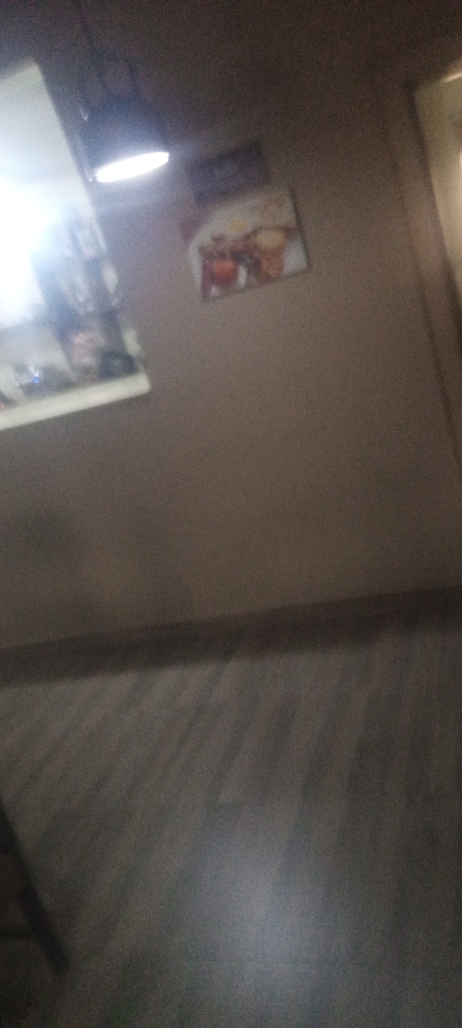 The image shows a blurred, indoor scene in what appears to be a restaurant or dining area. On the wall, there are two framed images: one is brightly lit and indistinct, possibly a menu or advertising; the other depicts food. The wall itself is a neutral, light color. The floor is a darker shade with a textured pattern, suggesting wood or tile. The room is dimly lit. The location is unknown, and there is no evidence of Kabala, Nigeria. There are no people visible in the frame.