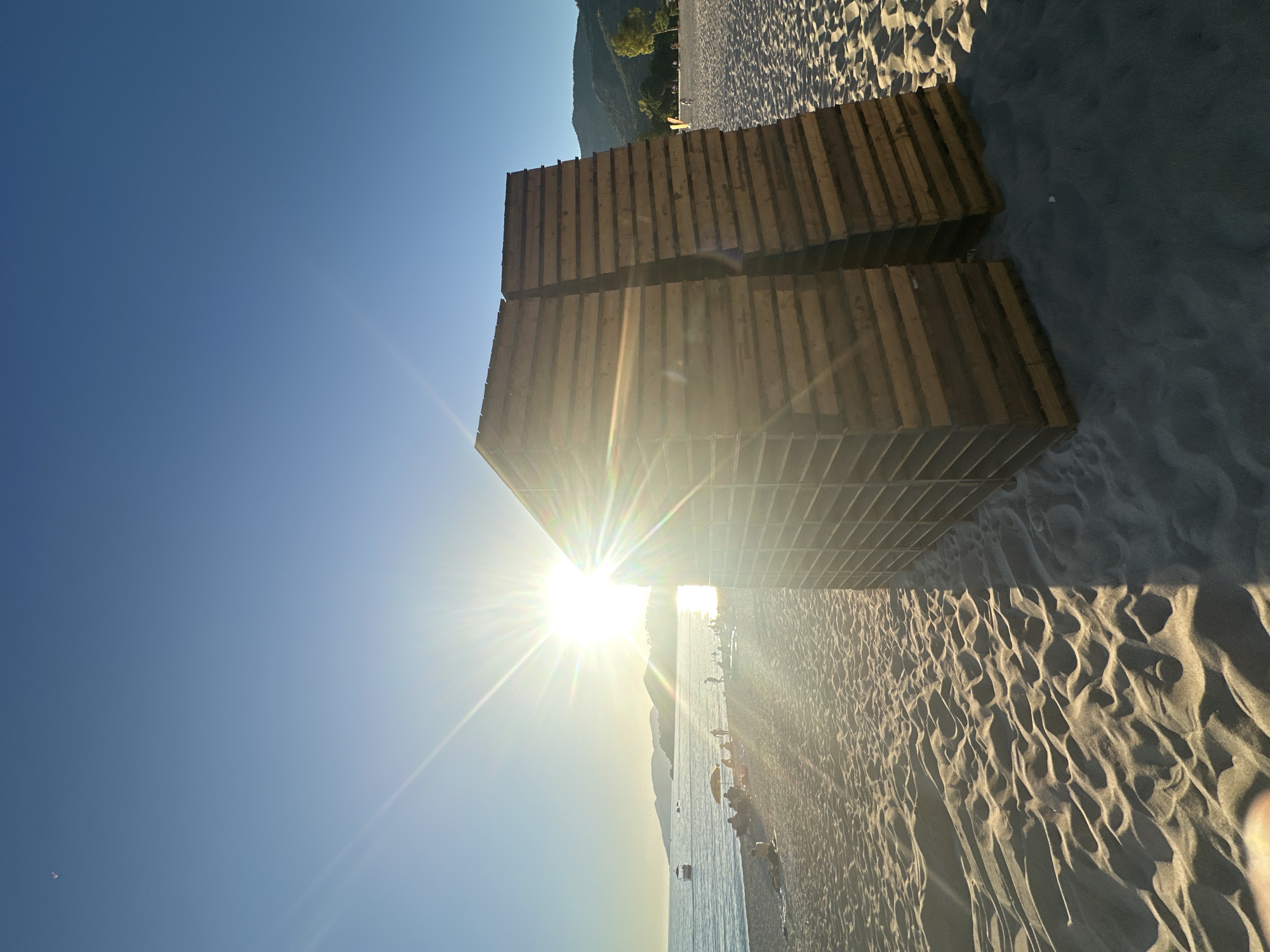 The image presents a vibrant, sun-drenched coastal scene in Fethiye, Turkey, captured with the camera pointing towards the bright sun, creating a prominent sunburst effect. Two large, dark wooden structures, possibly platforms or stacked pallets, are centrally located in the foreground, resting on the textured sandy beach.
Beyond these structures, the expansive sandy beach stretches out, showing distinctive ripples and patterns formed by wind or waves, with long shadows cast across its surface. The calm, clear blue sea occupies the mid-ground, reflecting the brilliant sunlight. In the distance, several small figures of people are visible, some on the beach and others in the water, suggesting leisure activities like swimming or sunbathing. A small boat is also discernible further out in the sea.
The background is dominated by a majestic range of mountains or hills, covered in lush greenery, that meet the clear horizon. The sky is a vivid, cloudless blue, gradually lightening towards the sun. A very small, distant object, possibly a kite or paraglider, can be faintly seen high in the sky to the upper left.
The bright, low-angled sun and the long shadows indicate either early morning or late afternoon, creating a warm and inviting atmosphere. The overall scene conveys a sense of peaceful relaxation and natural beauty. There is no visible text in the image.