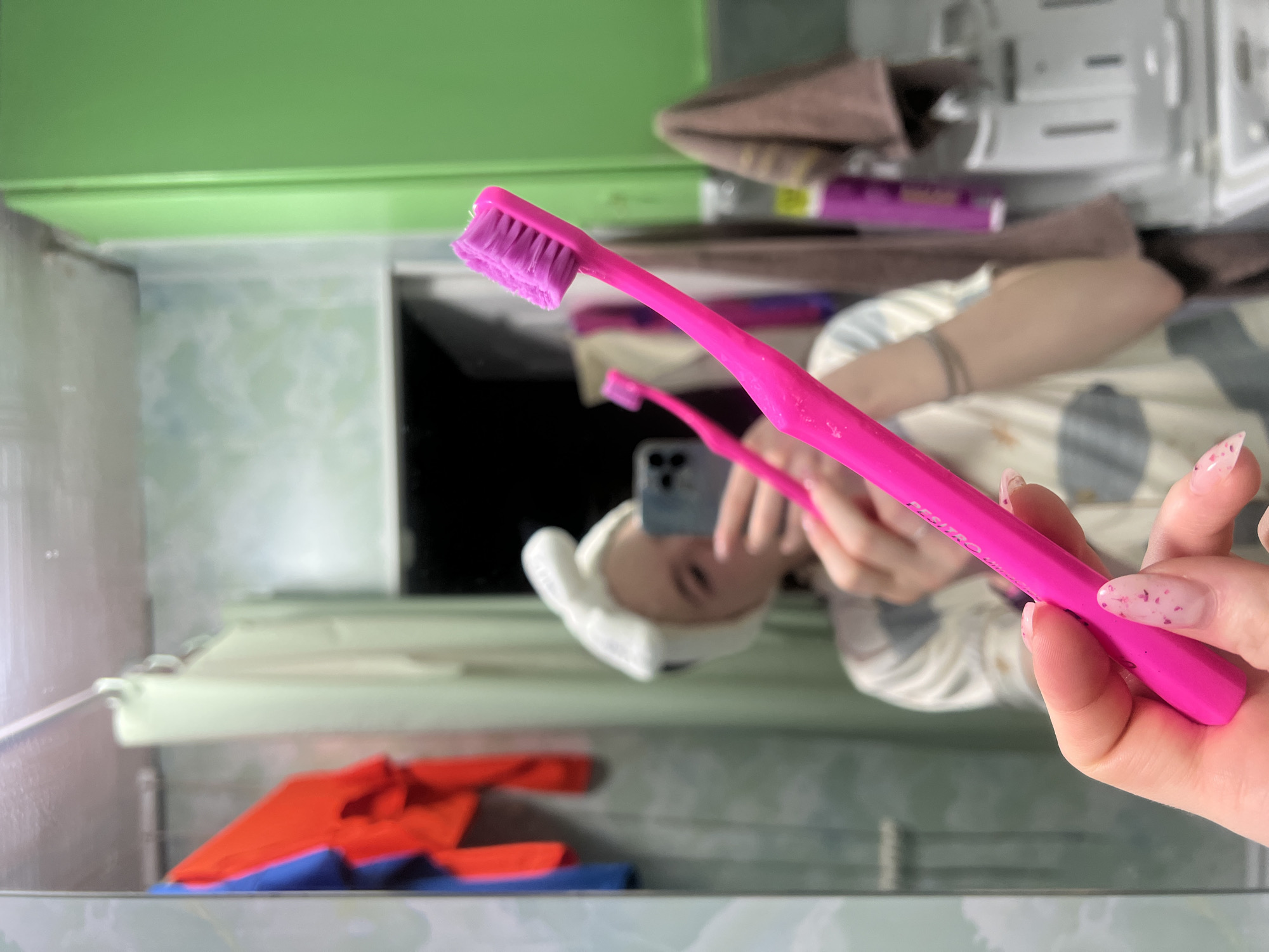 This media file captures a mirror selfie taken in a bathroom, with the original image rotated 90 degrees counter-clockwise.
The primary subject is a bright pink toothbrush with purple bristles, held in the foreground by a hand. The hand has light pink nail polish with small, dark pink/purple flakes. Faint white text is visible on the toothbrush handle, appearing to read "DENSITRO P.M." or similar.
The mirror reflects the person taking the selfie. This individual, whose head is wrapped in a white towel, is holding a smartphone (likely an iPhone due to its camera array) to capture the image. They are wearing a light-colored, patterned top. Due to the image's rotation, the reflected person's head appears tilted sideways.
The scene is a bathroom, evidenced by the large mirror, toiletries, and towels. Above the reflection, a vibrant green wall is visible, bordering a white shelf or counter. On this surface, various blurred bathroom items, including a purple container and folded brown towels, can be seen. Below the mirror, an orange towel and a blue towel are placed.
The activity taking place is a casual mirror selfie, possibly during a daily hygiene routine. No specific time of day or emotions are explicitly conveyed. The location context provided for this image is Chaikovsky, Russia.