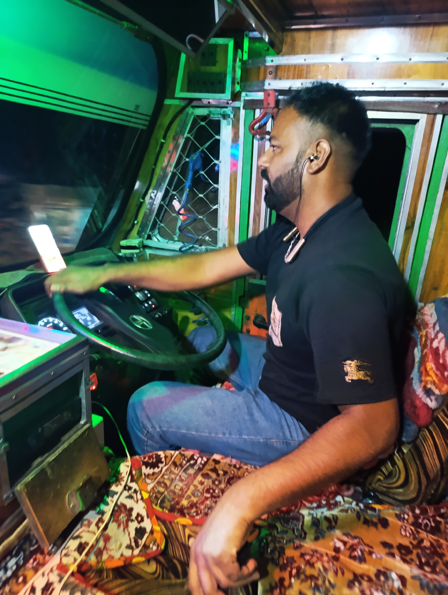 An Indian man with a beard and a fade haircut is driving a truck at night. He is wearing a black t-shirt and blue jeans. He has earbuds in and a necklace with a large pendant around his neck. The truck's interior is lit with green lights. The dashboard has a digital display and various buttons. The seats are covered in colorful, patterned fabric. The image does not provide enough information to determine the specific location in Gemur, India.