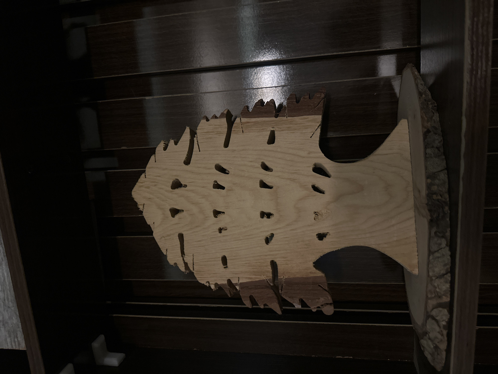 A wooden fish-shaped object is displayed on a dark, horizontal slatted surface, likely a shelf or wall. The fish is crafted from light-colored wood and has numerous small, oval-shaped holes cut into its body. The edges of the fish are rough and irregular, giving it a rustic appearance. A larger piece of wood with a natural, bark-covered edge is attached to the right side of the fish, extending outward like a tail or fin. The background consists of dark, horizontally oriented wooden slats that appear to be polished and slightly reflective, suggesting an indoor setting. The lighting is dim, casting shadows and highlighting the texture of the wood. The overall scene evokes a sense of handcrafted decor, possibly for a home or a workshop. No people, text, or specific weather conditions are visible. The location context of Pakuro, Nigeria, does not provide any directly observable cues within the image itself.