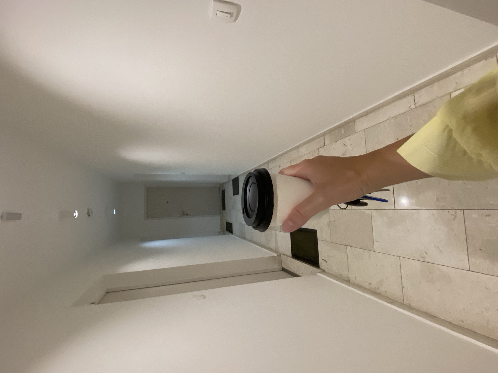 A person wearing a yellow shirt is standing in a narrow hallway holding a white and black cylindrical object. The hallway has white walls and a tile floor. There are several lights on the ceiling and a doorway at the end of the hallway. The location appears to be indoors. There is no information to confirm the location is in the city of Budva, Montenegro.