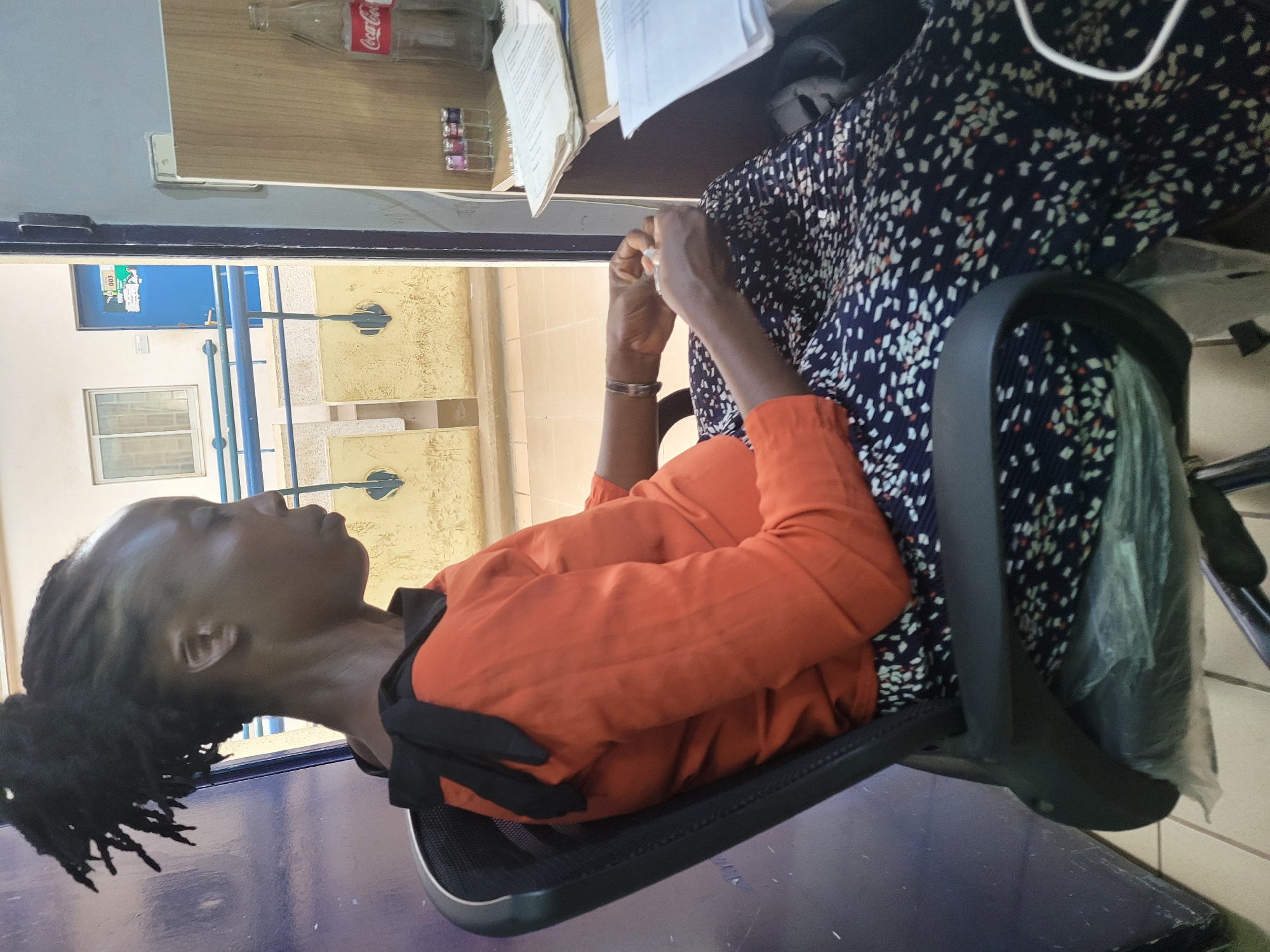 This image, rotated counter-clockwise, captures an indoor scene, likely an office or waiting area in Abuja, Nigeria, during the daytime. The primary subject is a person, appearing to be a woman, with dark skin and dreadlocks, sitting in a black office chair. She wears an orange long-sleeved top and a dark blue skirt patterned with white and orange specks. Her hands are clasped in front of her, holding a small white object, and she has a silver bracelet on her left wrist. Her gaze is directed upwards or straight ahead, with a neutral expression.
To her right, a light wood-grained desk or counter holds a clear glass Coca-Cola bottle, some papers or documents, and a small glass container with several colorful items, possibly candies or pills. Behind the seated person and through an opening or window, an outdoor or semi-outdoor area is visible. This area features a blue metal railing, a yellowish textured wall, and a white-framed window. Further in the background, a bright blue door is seen with a white sign displaying the text "003" and a graphic. The floor is tiled with light-colored squares. The overall environment suggests a functional or institutional setting.