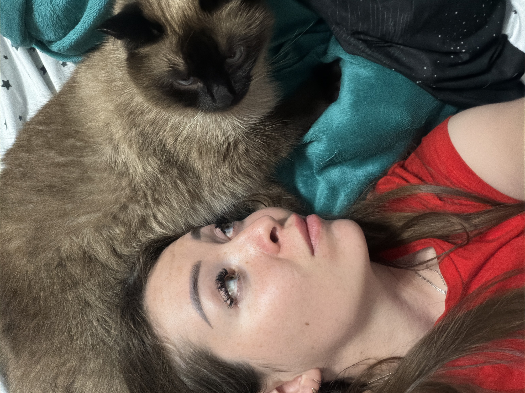 A close-up, high-angle shot shows a woman and a Siamese cat lying on a bed. The woman is on the left, facing right, with her head tilted up. She has long brown hair, is wearing a red t-shirt, and her left ear is adorned with a gold hoop earring. A silver necklace rests on her collarbone. Her expression is contemplative as she gazes upwards. Beside her, partially obscuring her, is a Siamese cat, its head facing forward. The cat has brown fur with darker points on its face and ears. It is nestled on a teal blanket. The background shows a white sheet with black star patterns and a dark fabric with white speckles. The scene appears to be indoors, in a bedroom, and there is no indication of the location being in the city of Япрыково, Russia.