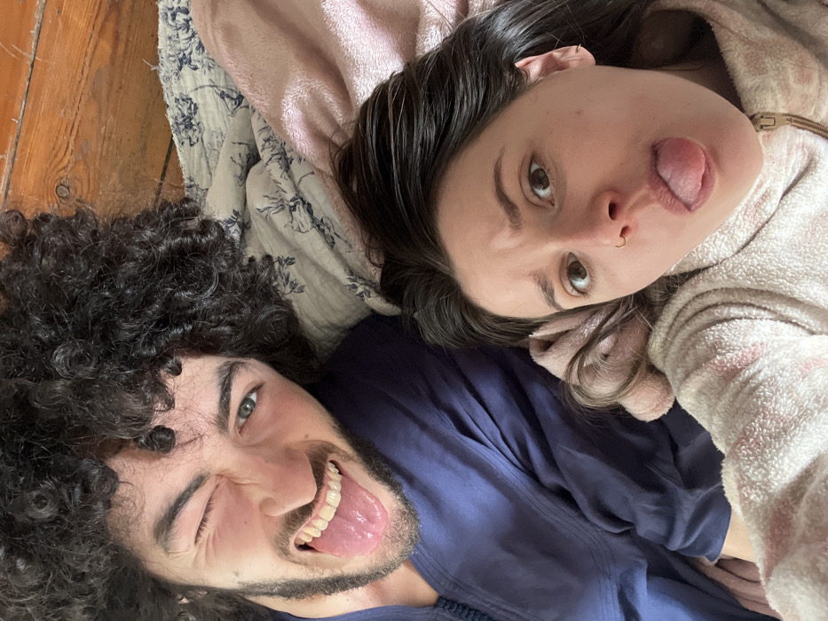 The image displays two individuals, one male and one female, lying on a bed in Carnin, France. The male, positioned on the upper left, has dark curly hair and a beard. He is wearing a dark blue garment with visible stitching on the collar. His right eye is winked shut, his mouth is open revealing teeth, and his tongue is extended. The female, positioned on the lower right, has dark straight hair and is wearing a light-colored, possibly pink or beige, hooded zippered garment with a striped texture. She is also extending her tongue, with her eyebrows slightly furrowed. A nose ring is visible on her left nostril and an earring on her left ear. Both individuals are looking directly at the camera. The bedding beneath them includes a patterned fabric with dark floral motifs and a solid light-pink textured blanket. A wooden surface, consistent with a headboard or wall panel, is visible in the upper right portion of the frame.