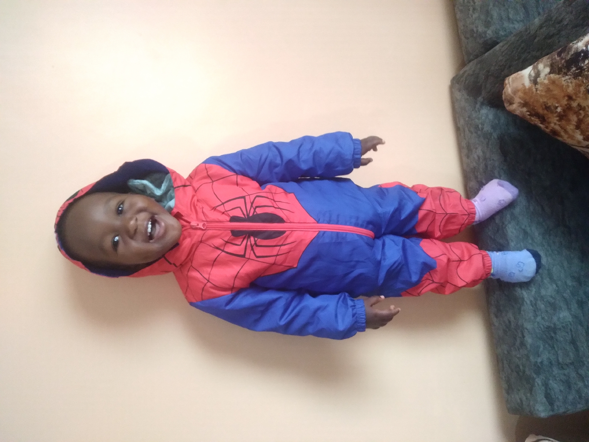 In Nyeri, Kenya, a young child, likely a toddler, stands on a dark green couch in front of a plain, peachy-orange wall. The child is wearing a full-body costume resembling Spider-Man's suit, with blue sleeves and legs, and a red torso adorned with a black spiderweb design and a spider emblem. The hood of the costume is up, revealing the child's joyful expression, with wide-open mouth and bright eyes. The child is also wearing light blue socks on their left foot and light purple socks on their right foot. The lighting suggests an indoor setting, possibly a living room, with the wall serving as a backdrop.