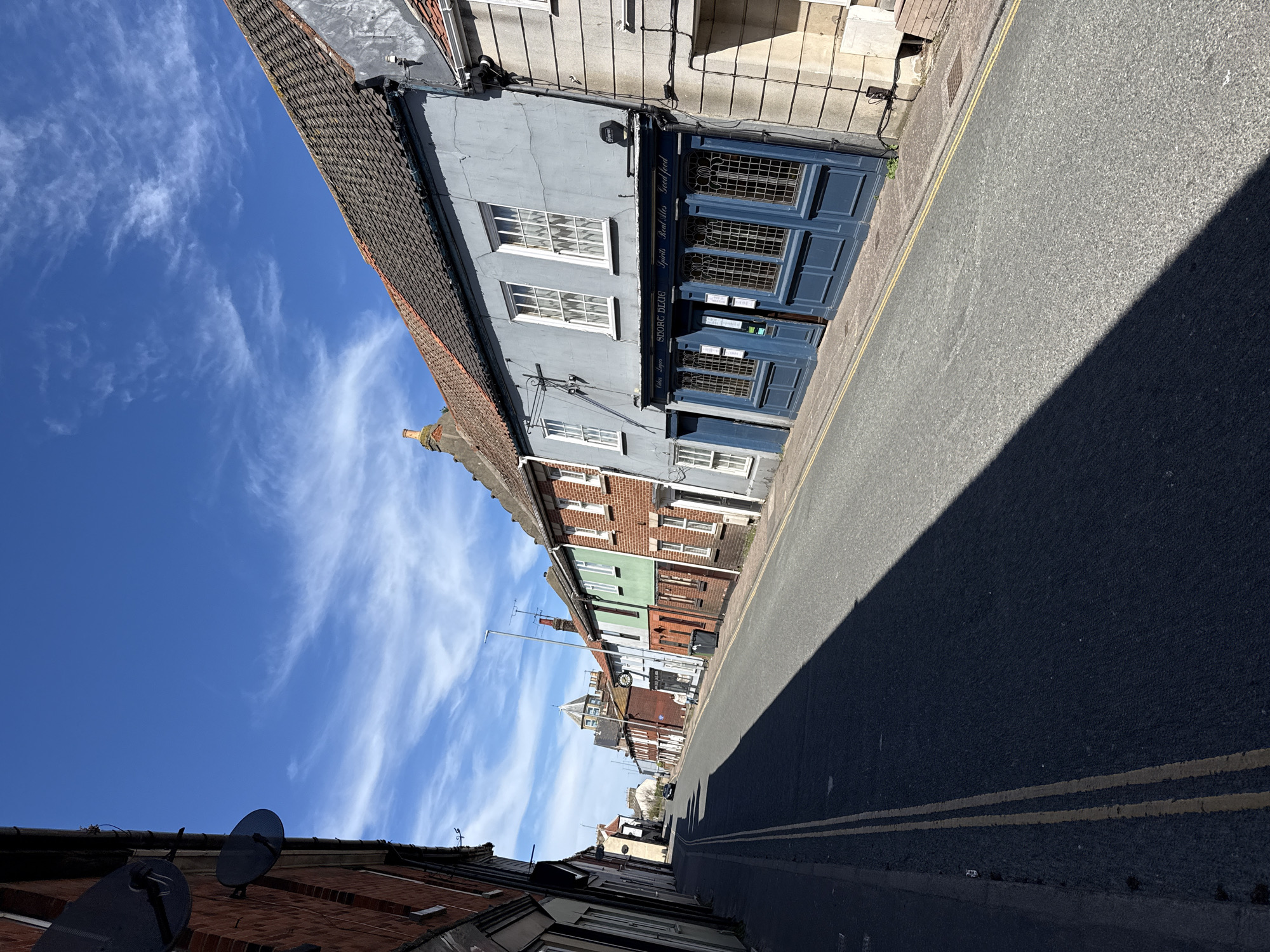 This media file presents a sunlit street scene in Gorleston-on-Sea, United Kingdom, captured from a slightly low perspective looking down a row of buildings. The clear, bright blue sky is adorned with scattered, wispy white clouds, indicating fair weather. Long, sharp shadows stretch across the asphalt road, suggesting a time of day with a low sun, either morning or late afternoon.
On the left side of the frame, a succession of terraced buildings lines the street. In the foreground, a red brick building with two black satellite dishes on its roof is visible. Adjacent to it stands a prominent light blue-grey building featuring white-framed windows and a dark blue shopfront. This establishment displays clear text on its fascia: "GORD BLUES" and "Open Late" on the left, and "Spirits," "Real Ales," "Good Food" on the right. Further down the street, a variety of colorful buildings, including green and red brick facades, are visible, with a distinctive building featuring a clock face in the mid-distance.
The street itself is an asphalt road with double yellow lines running parallel to the curb on the left. A significant portion of the road on the right is covered by a deep, dark shadow, contrasting with the sunlit section and highlighting the intensity of the sunlight. No people or specific activities are visible, lending a quiet atmosphere to the street. The architectural style of the buildings is characteristic of a traditional British town or coastal area.