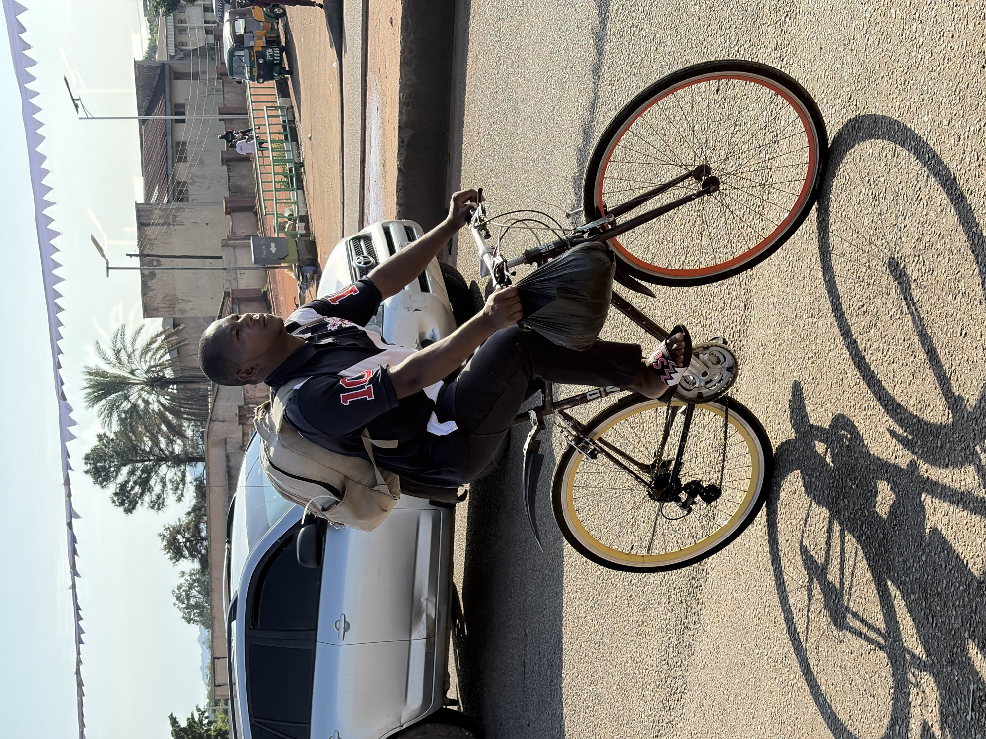 A young Black male is riding a bicycle on a paved road in Gura Top, Nigeria. He is wearing a black polo shirt with a white collar and a red number "10" on the sleeve, dark pants, and sandals. He has a light-colored backpack on his shoulders and is carrying a black plastic bag in his left hand. The bicycle has a black frame, a rear fender, and wheels with yellow and orange rims. The sun is casting a long shadow of the bicycle and rider on the road. In the background, there are vehicles, including a silver SUV and a yellow and green auto-rickshaw, as well as buildings and trees under a bright sky.
