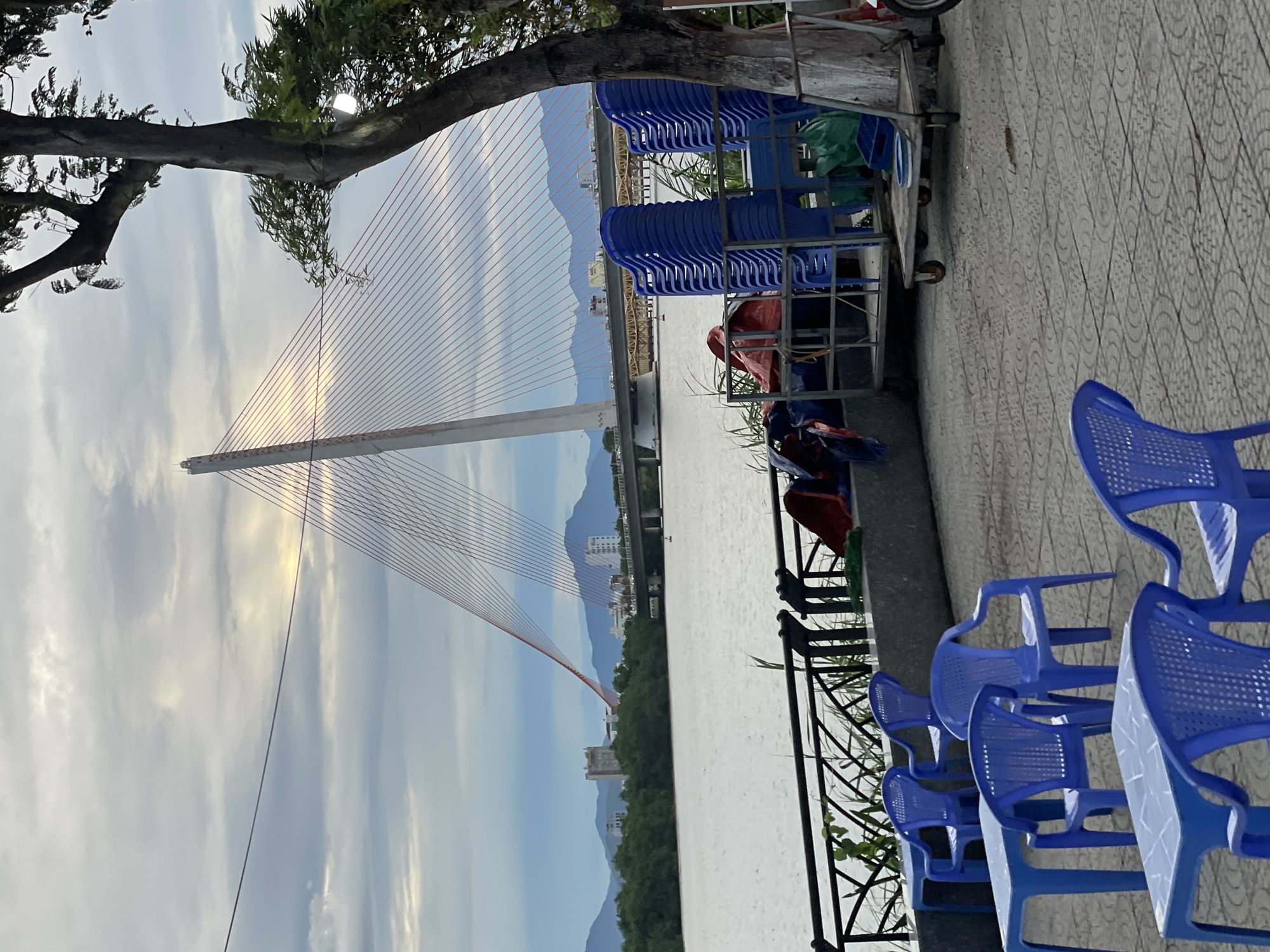The image depicts a waterfront scene in Hoi An, Vietnam, likely along the Thu Bon River. In the foreground, several blue plastic chairs and tables are arranged on a paved area. Behind them, a stack of similar blue chairs is visible, along with a metal cart holding more chairs and some tarpaulin-covered items. A large tree with a textured trunk stands to the right, partially obscuring a lamp attached to it.
The midground features a body of water, likely a river, with a calm surface reflecting the overcast sky. A black metal railing runs along the edge of the waterfront. Beyond the water, a large, modern suspension bridge dominates the landscape, characterized by its tall white pylon and numerous red suspension cables. Several buildings and dense green foliage line the far bank of the river. In the distance, mountains are visible under a cloudy sky. The overall atmosphere suggests a slightly overcast day, possibly late afternoon or early morning, with the lighting diffused by clouds. No people are clearly visible in the scene.