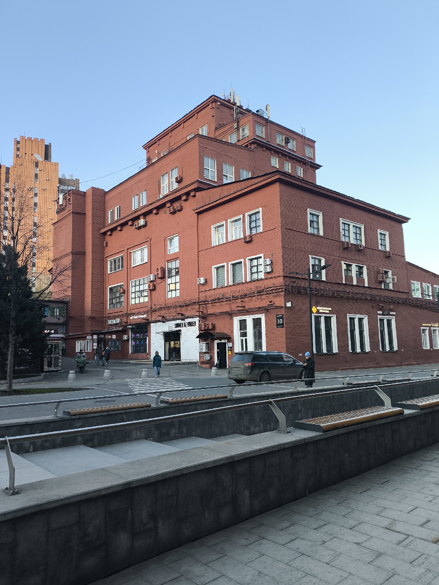 This image captures a vibrant street scene in what appears to be a European city, with context indicating Moscow, Russia. The focal point is a large, multi-story building constructed of red brick, featuring a distinctive stepped or terraced upper structure with several roof levels. Numerous white-framed windows, many with air conditioning units beneath them, dot the facade. The building's architecture suggests a blend of functional and possibly industrial or early 20th-century design. Various antennae and satellite dishes are visible on its rooftops.
In the background to the left, a taller, lighter orange-brown building with many windows rises against a clear, light blue sky. A bare-branched tree stands in the foreground on the left, indicating autumn or winter.
At street level, the scene is bustling with activity. A paved road features a clearly marked pedestrian crossing. A dark-colored SUV is either parked or moving slowly near the sidewalk on the right. Several people are visible: one person in a blue hat and dark coat stands beside the car; a group of people walks on the sidewalk further down; and another individual pushes a green cart. A pedestrian is also seen crossing the street in the distance.
The foreground features modern urban landscaping elements, including large concrete structures that serve as planters or steps, with integrated wooden benches and sleek metal railings.
Visible text on the red brick building includes the number "10" near a corner. Shop signs display names such as "SOMMER AF VINDEN," "INTERNET DYNAMICS," and partially visible "SOVREMENNYY MEBEL" (likely "Modern Furniture" in Russian). A small kiosk or booth has "КАФЕ" (Cafe) written on it. The time of day is clear, bright daylight, indicated by the crisp lighting and the blue sky.