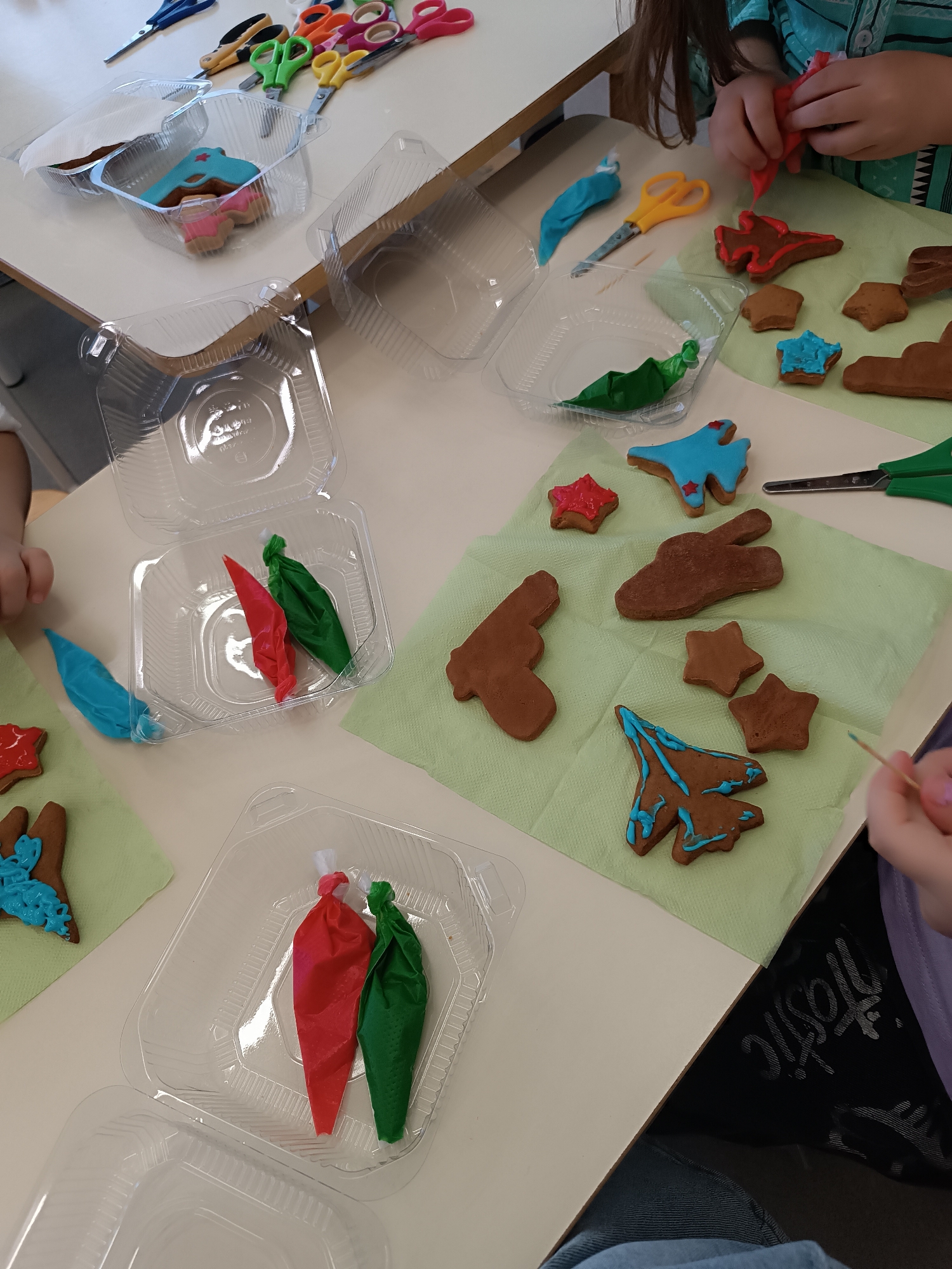 Several children are decorating cookies shaped like stars, airplanes, guns, and clouds on a light-colored table covered with light-green napkins. There are plastic containers nearby holding tubes of red, green, and blue icing, as well as some decorated cookies. There are also multiple pairs of scissors on the table. The location is unknown.