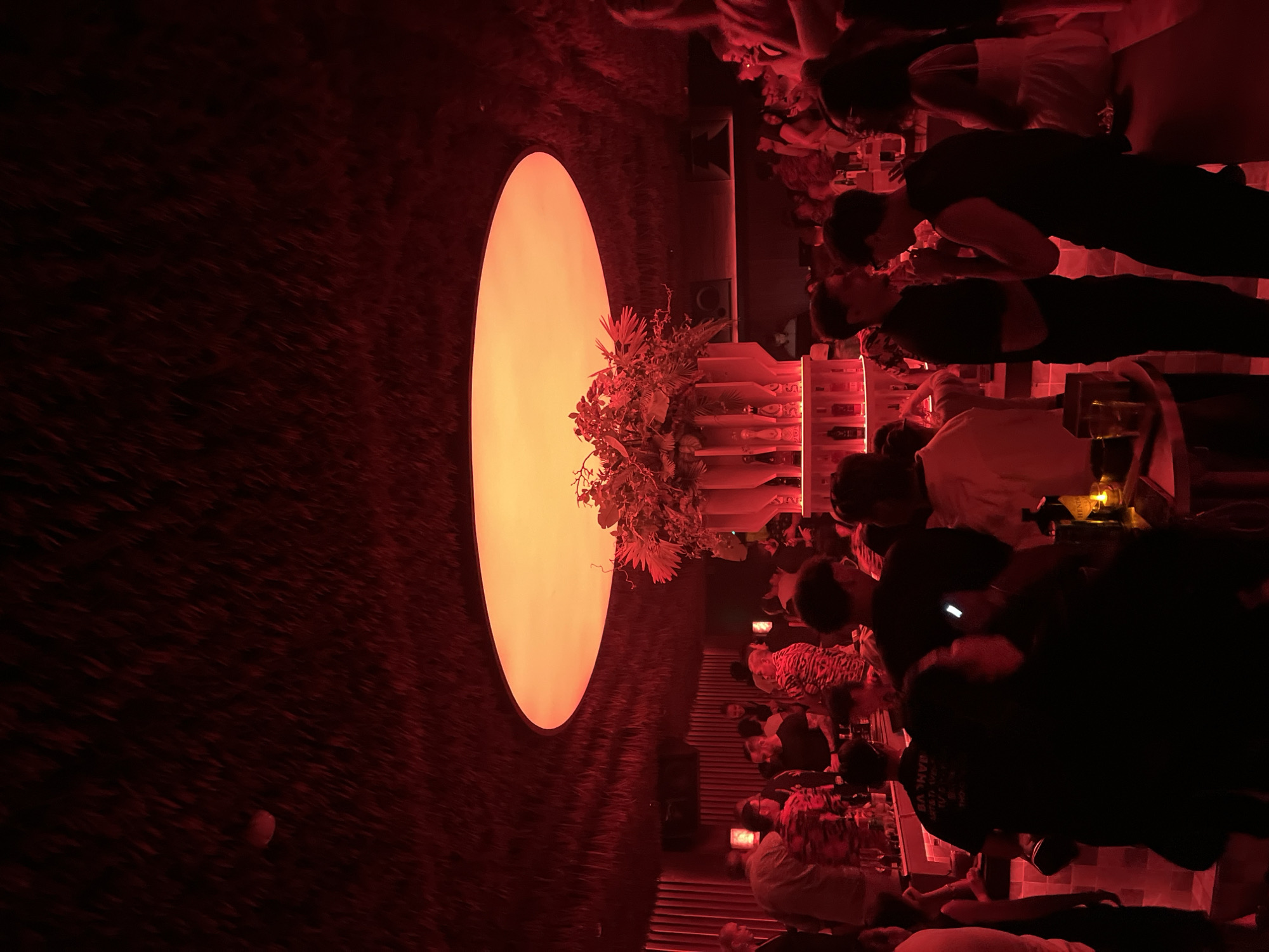In Thủ Đức, Vietnam, a dimly lit interior space, possibly a bar or lounge, features a large, oval-shaped light source emitting an orange glow against a dark, textured wall. Plants are arranged around a tiered shelf unit with bottles and glassware. A group of people are gathered at tables, some standing and some seated. Many people are engaged in conversations and interactions. The space is lit with red and orange lighting.