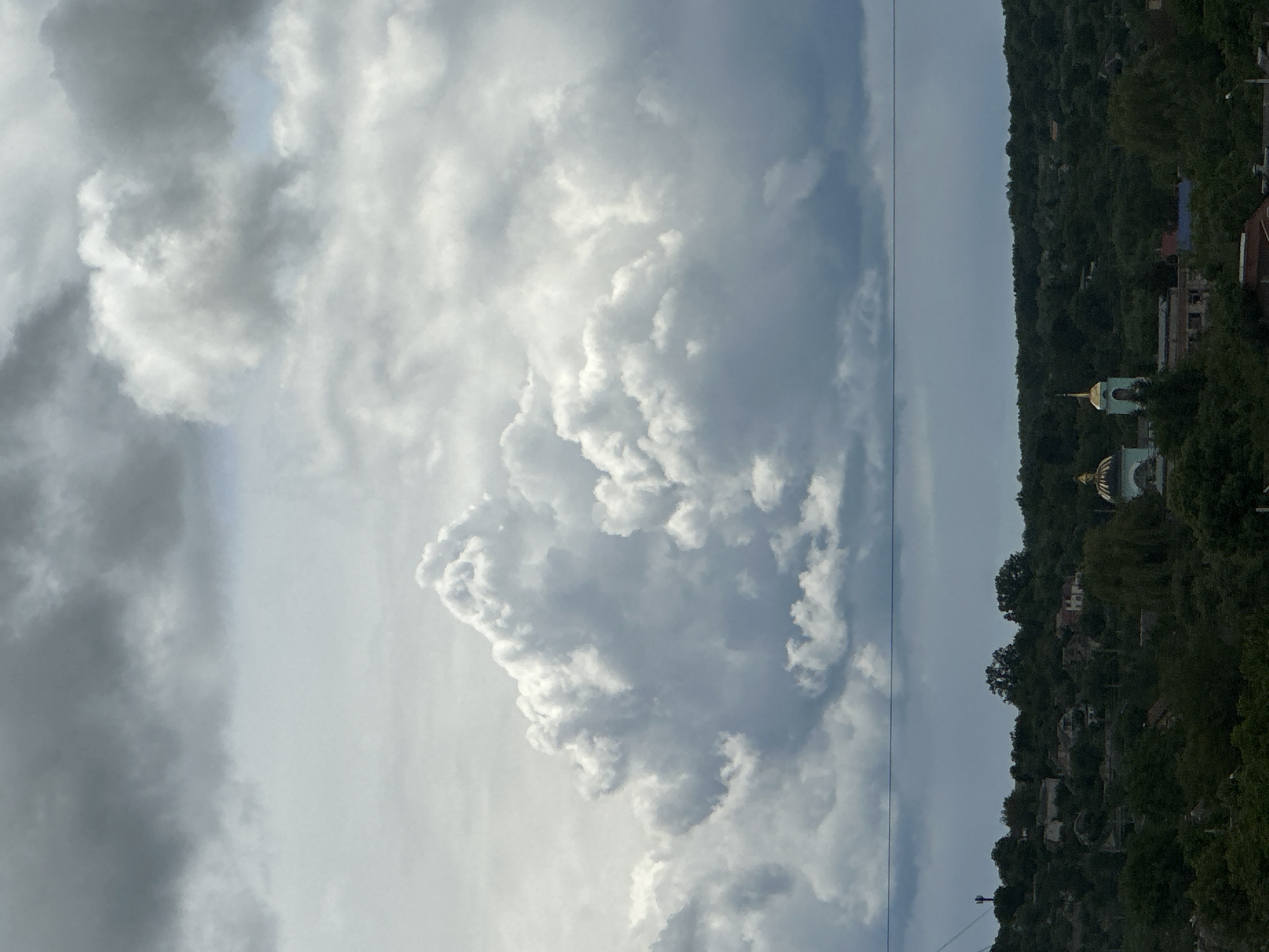 A view of the sky with storm clouds is oriented sideways. On the right side of the frame, a hillside covered in dense trees is visible. A church with golden domes is nestled among the trees. Houses dot the hillside. A power line runs vertically through the image near the edge of the hillside. The location is Unknown, Unknown.