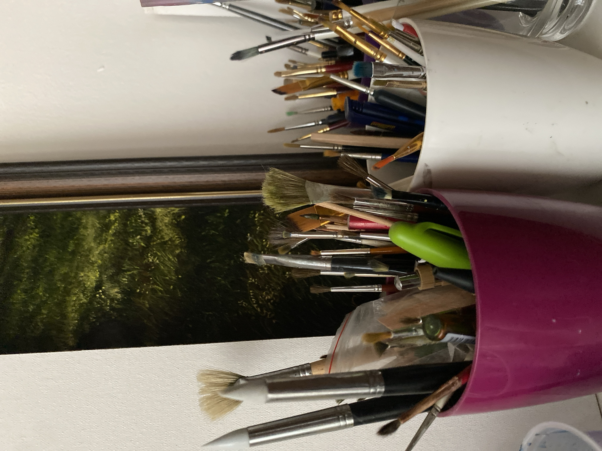 This is a close-up, eye-level, interior photograph of an artist's workstation. The scene is located in Bílina, Czechia. Two open-top containers, a magenta-colored one on the left and a white one on the right, are filled with a collection of art brushes of various sizes and types. Some brushes are positioned upright, while others are angled. In the magenta container, a bright green pen is visible among the brushes. Behind the containers, a framed artwork depicting a grassy field with hints of sunlight is partially visible, leaning against a white wall. The lighting is moderate, with some shadows indicating a natural or diffused light source. The overall impression is one of an organized, functional art studio space.