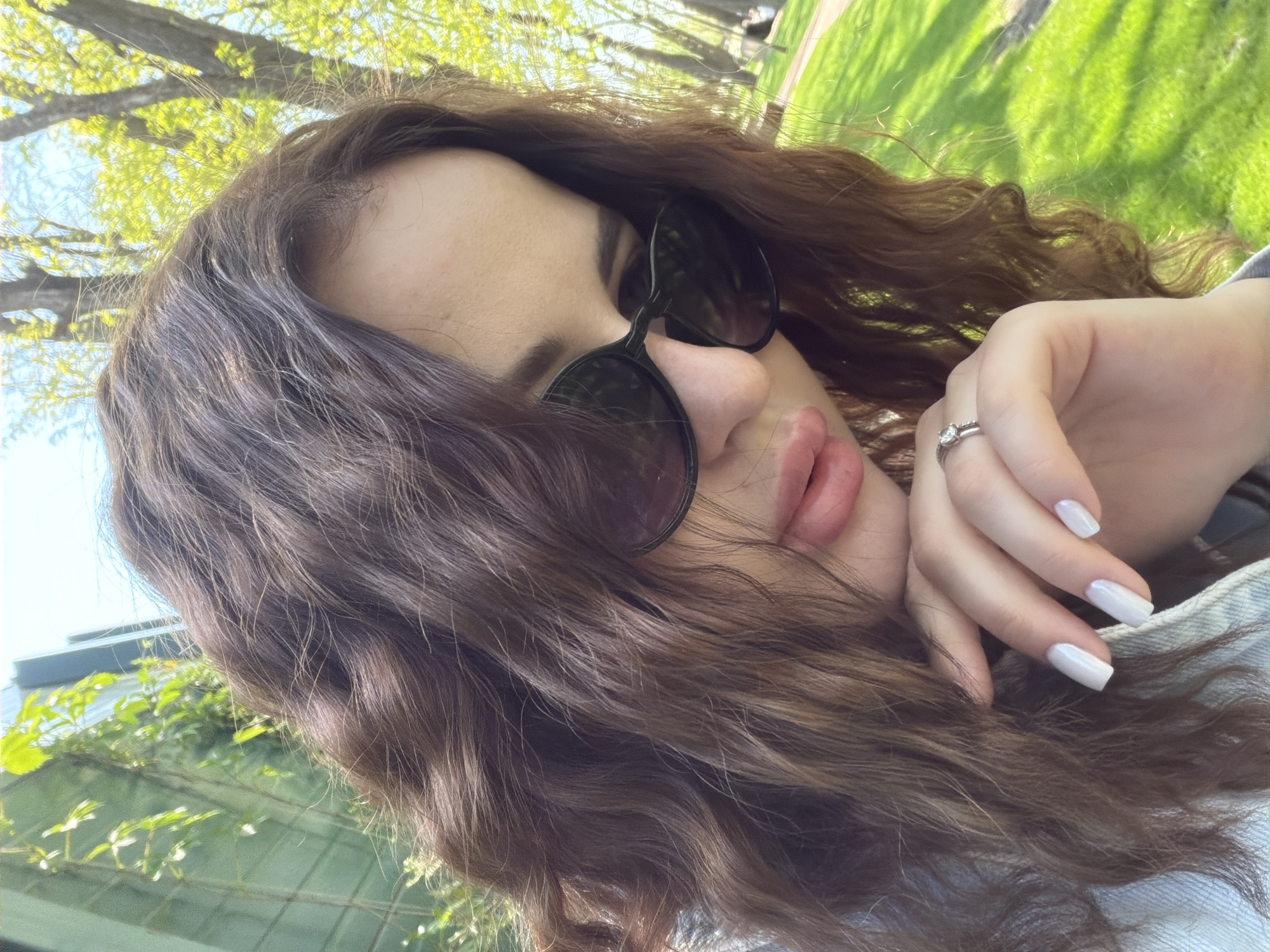 A medium shot shows a woman with light skin and brown, wavy hair wearing sunglasses. She is wearing a ring on her left ring finger, and her fingernails are painted white. She is outside, surrounded by foliage and trees. The location is unknown.
