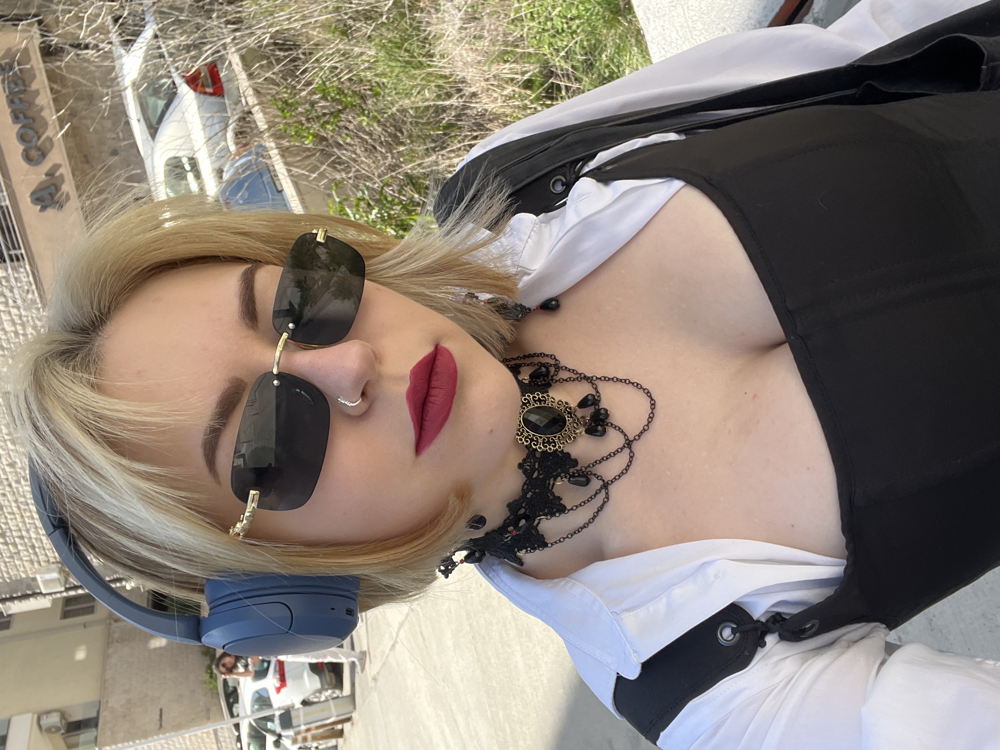 The image shows a woman with blonde hair wearing sunglasses and blue headphones. She has a nose ring and is wearing dark red lipstick. She is also wearing a white shirt with a black corset-like garment on top, as well as a black necklace with a decorative center. The background features blurred elements of a city, including buildings and a car. Based on the sign in the background, the location is Boreti, Montenegro.