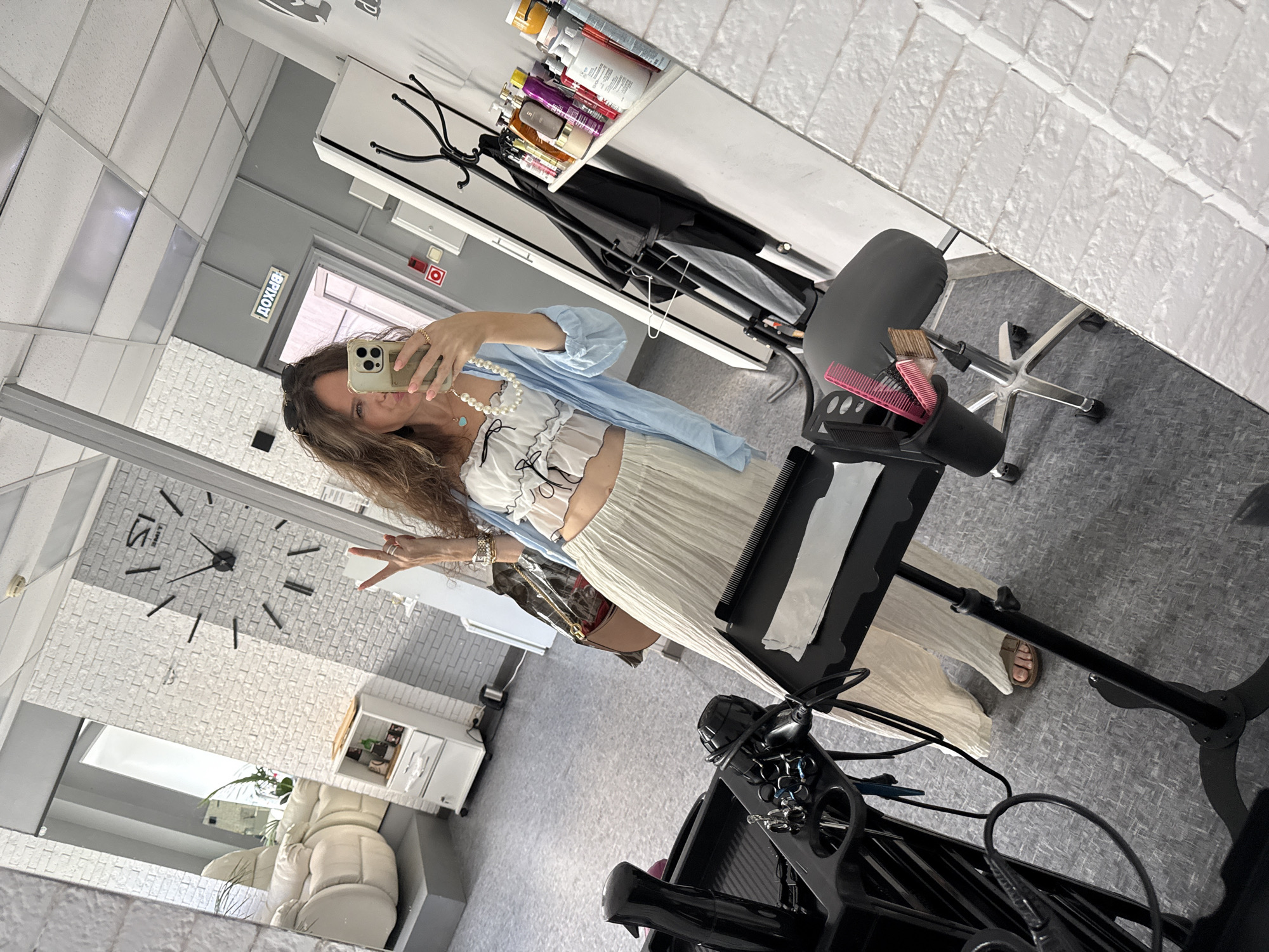 A woman is taking a mirror selfie in what appears to be a beauty salon in Moscow, Russia. The woman has long, wavy brown hair and is wearing a white crop top with black trim, a light blue button-down shirt, and a long, pleated cream-colored skirt. She is accessorized with a pearl necklace, rings, and a watch. She is holding a smartphone in her right hand, angled to capture the reflection. Her left hand is raised, with two fingers extended in a peace sign.
In the reflection, the salon environment is visible. To the left, there is a plush white chair and a white cabinet with drawers. On the wall behind the woman, a large, modern clock with Roman numerals is mounted. To the right, a shelf is laden with various bottles and containers, presumably hair and beauty products. A coat rack stands next to the shelf. A styling trolley with a hairdryer and combs is positioned in the foreground. The floor is carpeted with a grey, patterned material. A sign with Cyrillic lettering, "ВХОД," meaning "ENTRANCE," is visible above a doorway in the background. The walls are decorated with white brick-patterned tiles.
