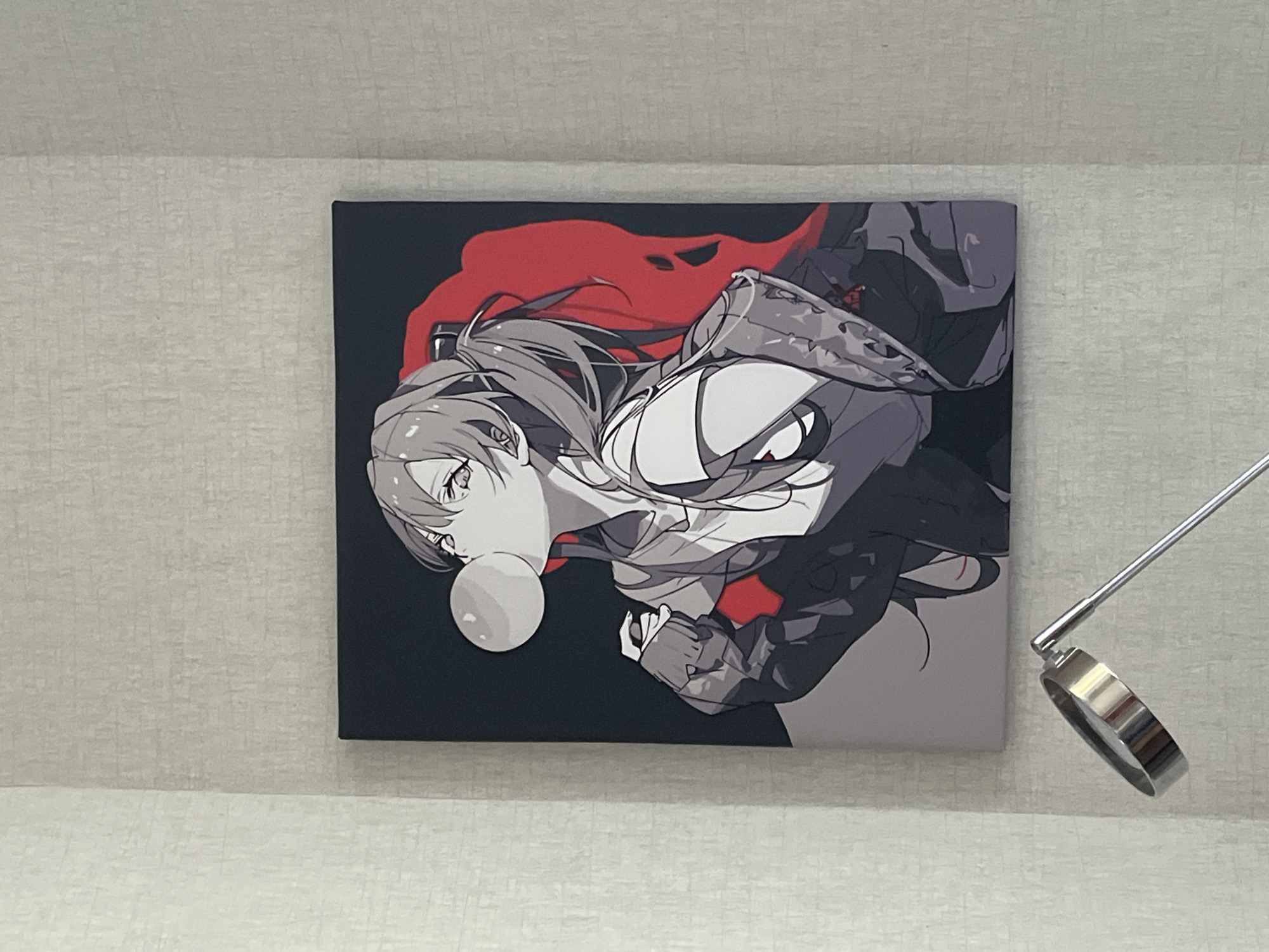 The image shows a framed anime-style artwork hanging on a textured, light-colored wall. The artwork depicts a young woman with her hair in a ponytail, blowing a large bubblegum bubble. She is wearing a casual outfit consisting of a t-shirt and a jacket. A large red abstract shape is behind her. In the foreground, a portion of a silver metallic desk lamp is visible, with its circular head angled towards the artwork. The scene appears to be indoors, likely in a home or office. There is no identifiable information about the location being in Minsk, Belarus.