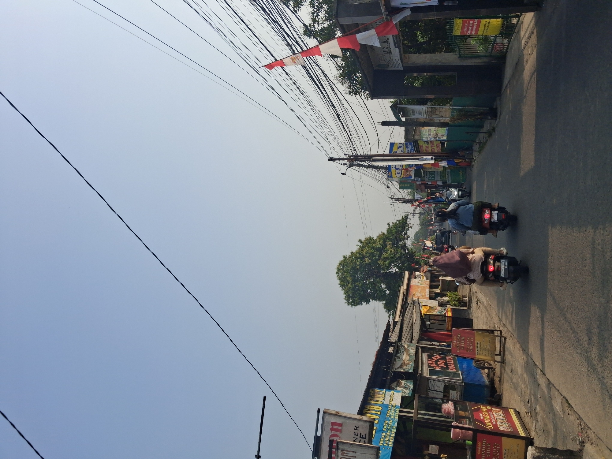 The image shows a street scene in Bojong Gede, Indonesia, under a clear, bright sky. Power lines are strung across the sky, creating a dense network above the street. On the left side of the road, there are several small businesses and food stalls, indicated by signs such as "NER CE" and menus displayed outside. One stall appears to be selling "LONTONG SAYUR". Further down the road, there are more shops and some small trees.

On the right side of the road, there is a gated entrance with an Indonesian flag hanging from a pole. Above the entrance, a sign reads "MIS. AL". The gate is green, and a yellow banner with Indonesian text is attached to it.

Two people are riding on a single motorcycle in the foreground, traveling away from the viewer. The passenger wears a light brown hijab and a light-colored top, while the driver wears a dark top. Another motorcycle with two passengers is visible further ahead on the road. In the distance, other vehicles, including cars and motorcycles, are also present on the street. The overall environment suggests a moderately busy street in a residential or commercial area of Bojong Gede.