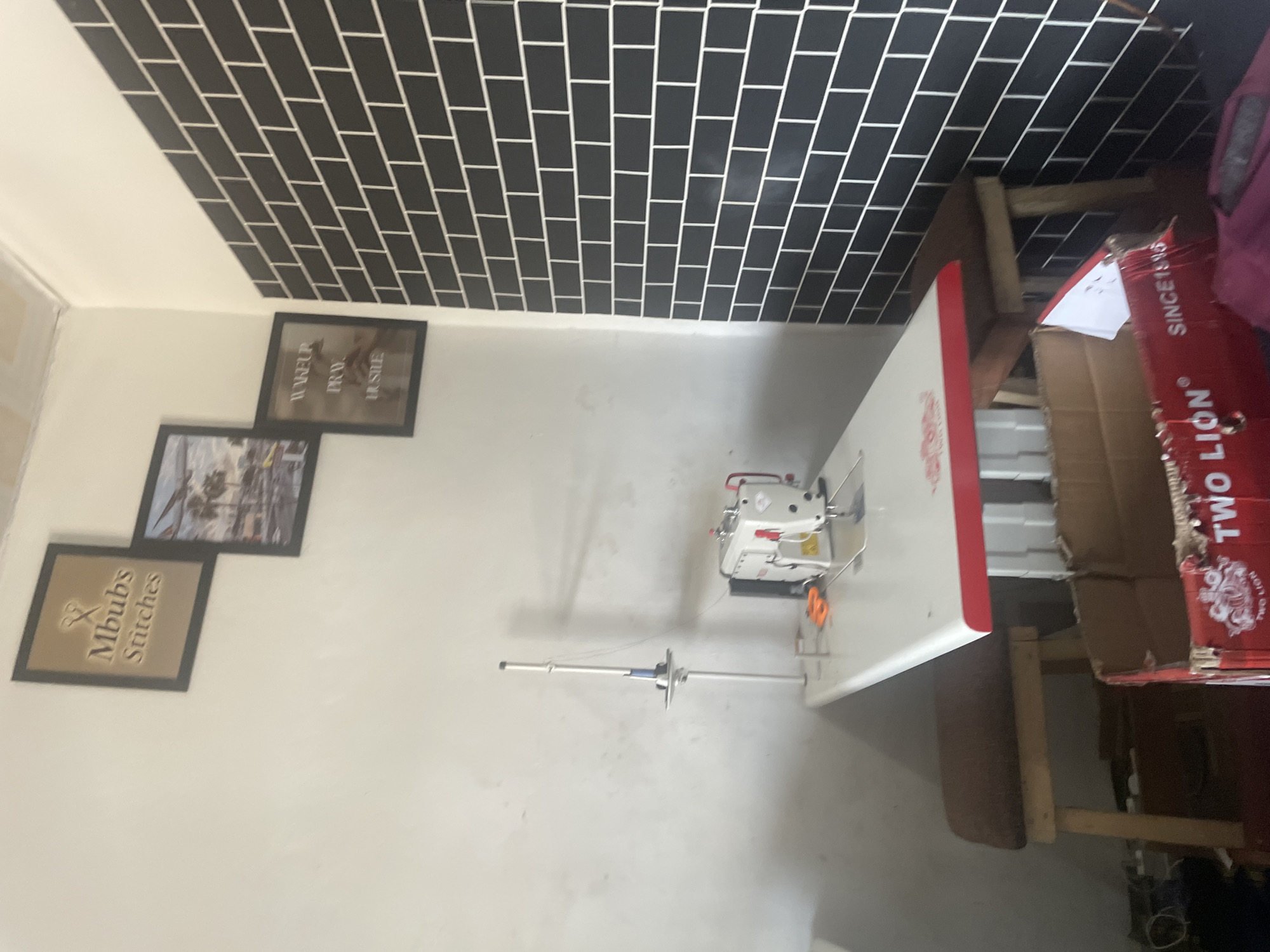 The image depicts a sewing room located in Jalingo, Nigeria. The room features a white sewing machine on a table with a red trim, situated against a wall adorned with black subway tiles. To the left of the sewing machine, a white wall is visible with three framed pictures hanging on it. The leftmost picture is a square frame containing the text "Mbubs Stitches" and a pair of scissors. To its right, a rectangular frame displays a scenic image of palm trees and buildings. The third frame, positioned below the scenic image, contains the text "WAKE UP. PRAY. HUSTLE." Two wooden stools with brown cushions are placed under the table. A cardboard box with red and white branding, including the words "TWO LION" and "SINCE 1918," is in the foreground. Various electrical cords and other miscellaneous items are present on the floor. The lighting suggests an indoor setting, likely during the daytime.