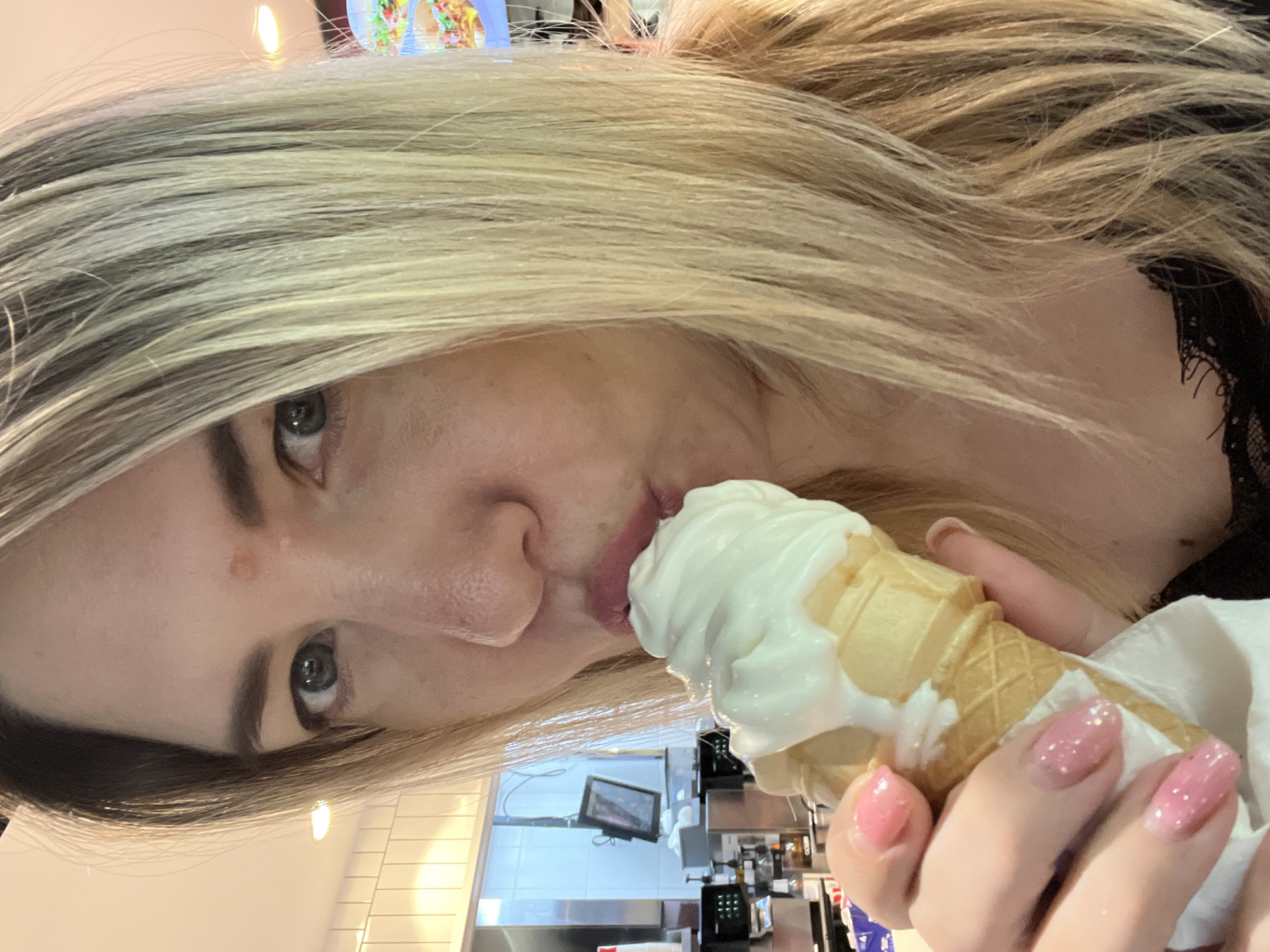 A woman with blonde hair is eating a vanilla soft-serve ice cream cone. She is holding the cone in her right hand and bringing it to her mouth. The background shows a brightly lit interior, possibly a fast-food restaurant or ice cream parlor, with white tiled walls and a counter area with various equipment. The woman has on a black lace top. The setting is not identifiable as Sochi, Russia.