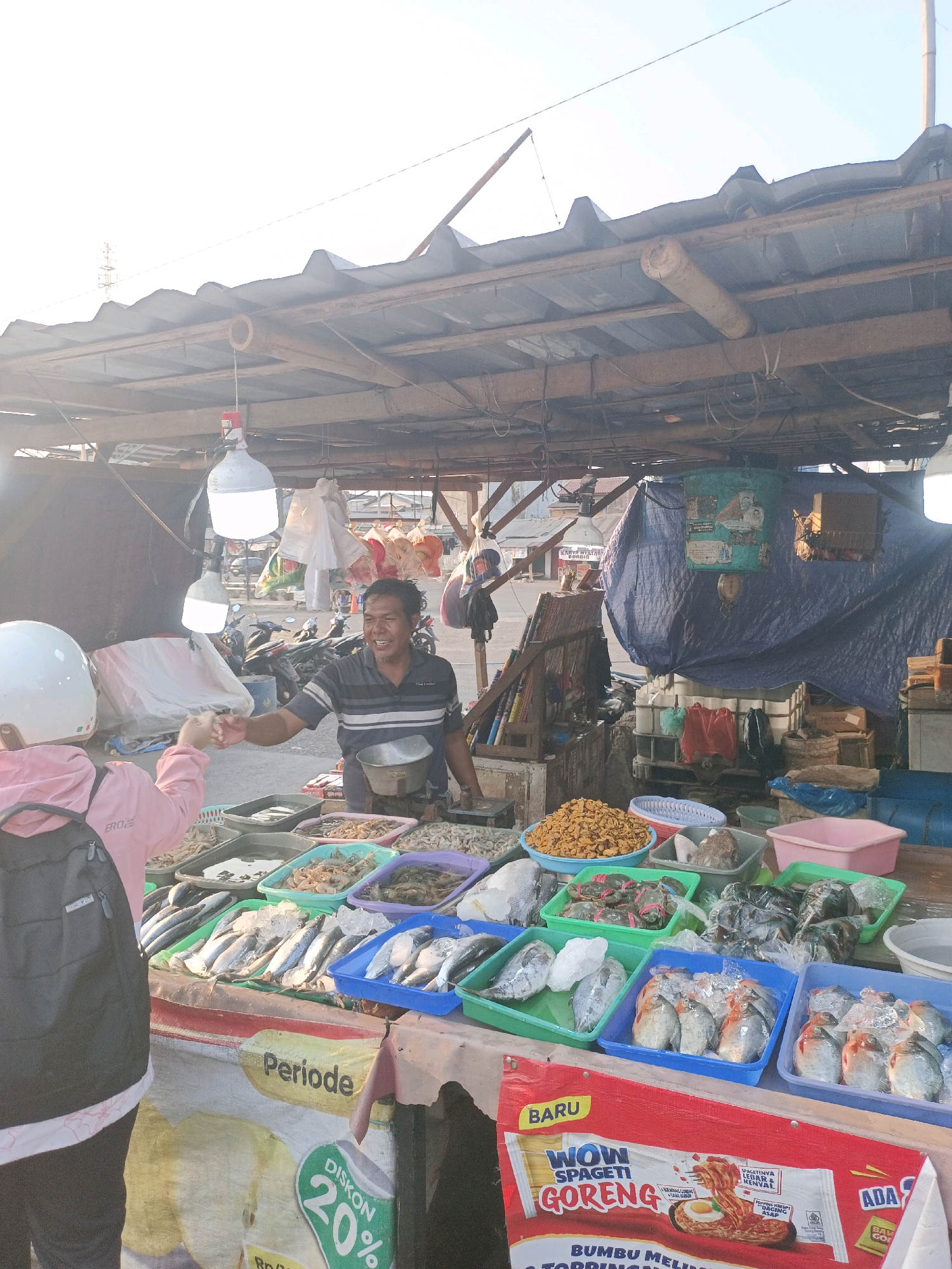 A medium shot captures a market stall in Karawang, Indonesia, under a corrugated tin roof supported by wooden beams. The stall displays a variety of fresh seafood, including fish, shrimp, and possibly crabs, arranged in plastic containers. A male vendor, wearing a striped t-shirt and smiling, is interacting with a customer from behind the stall. The customer, a young person wearing a pink hoodie and a white helmet, is partially visible from the side, reaching out to the vendor. The environment appears to be an outdoor market, with motorcycles and other stalls visible in the background. The lighting suggests it is either early morning or late afternoon. A large banner advertising "WOW SPAGETI GORENG" is prominently displayed at the bottom of the stall.