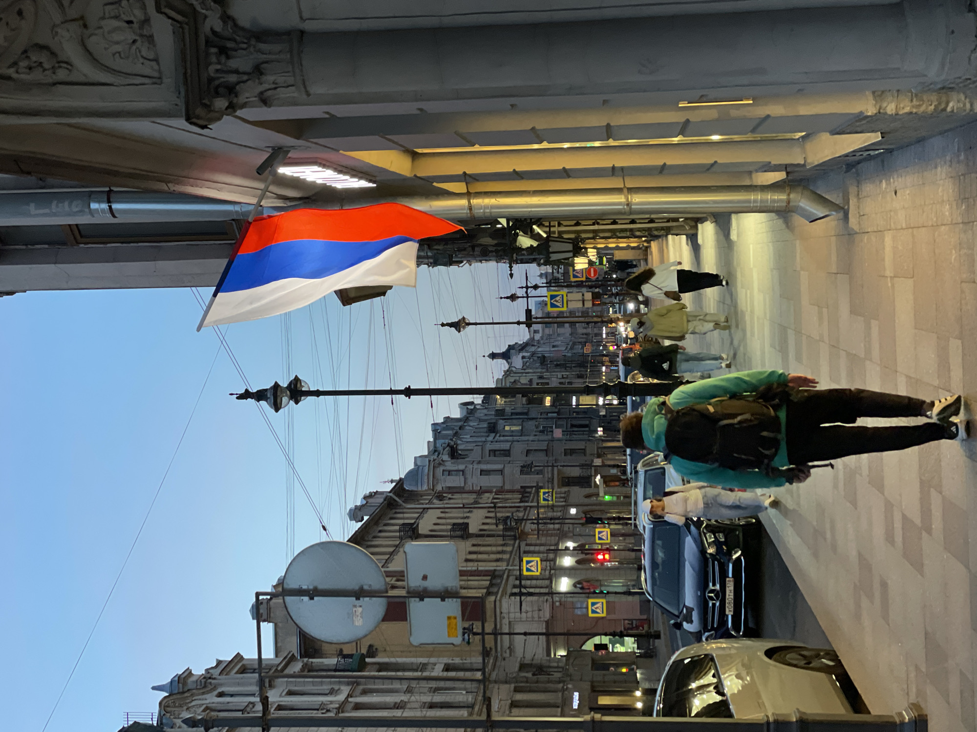 A street scene in Saint Petersburg, Russia. A red, blue, and white flag hangs beneath a portico. People walk on the sidewalk alongside parked cars. The street has traffic lights and pedestrian crossing signs. Buildings line the street. The sky is visible above the buildings.