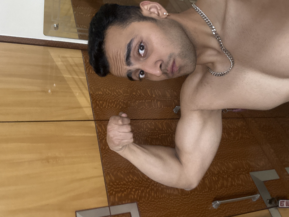 A shirtless man with dark hair poses with a flexed right arm. He has a chain necklace and an earring. He looks directly at the camera with a slight pout. The background consists of a glossy surface divided into two distinct areas: one is a light, wood-grained pattern, and the other has a darker, patterned texture. There are metallic or reflective accents on the glossy surface near the bottom. The image is oriented horizontally.