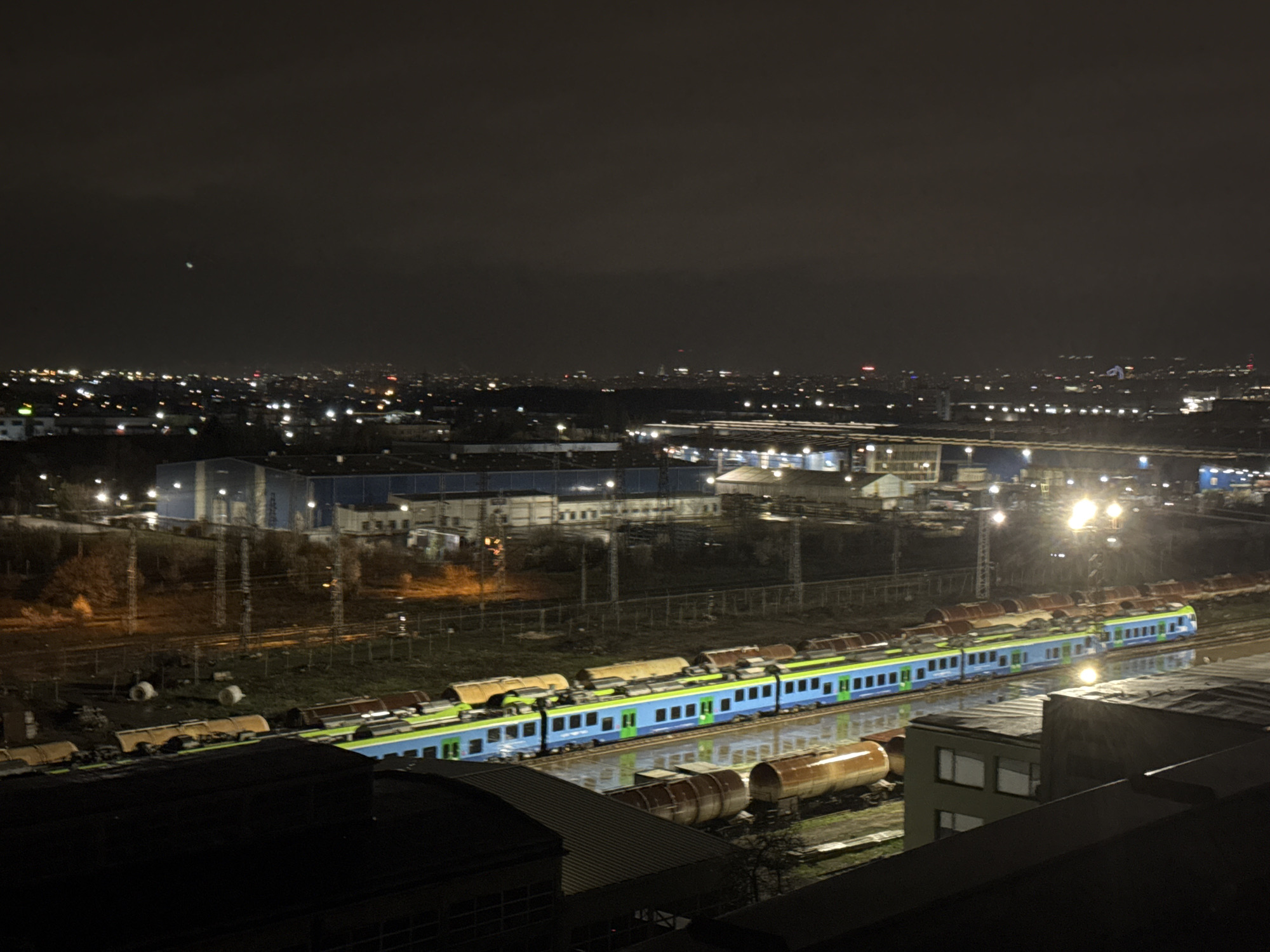 The image shows an elevated view of a train yard at night, likely within or on the outskirts of a city. Distant city lights illuminate the horizon, suggesting an urban setting.
A train with blue and light green coloring sits on the tracks. There are several tank cars nearby. In the foreground, parts of buildings with dark rooftops are visible. There are streetlights and buildings of industrial character on the other side of the rail yard.
It is likely that the scene is located in Sofia, Bulgaria due to the presence of BDZ trains, the national railway company of Bulgaria, which utilizes this color combination for their vehicles. Also, the scene with industrial yards on the outskirts of a city in this view fits in with the outskirts of Sofia.