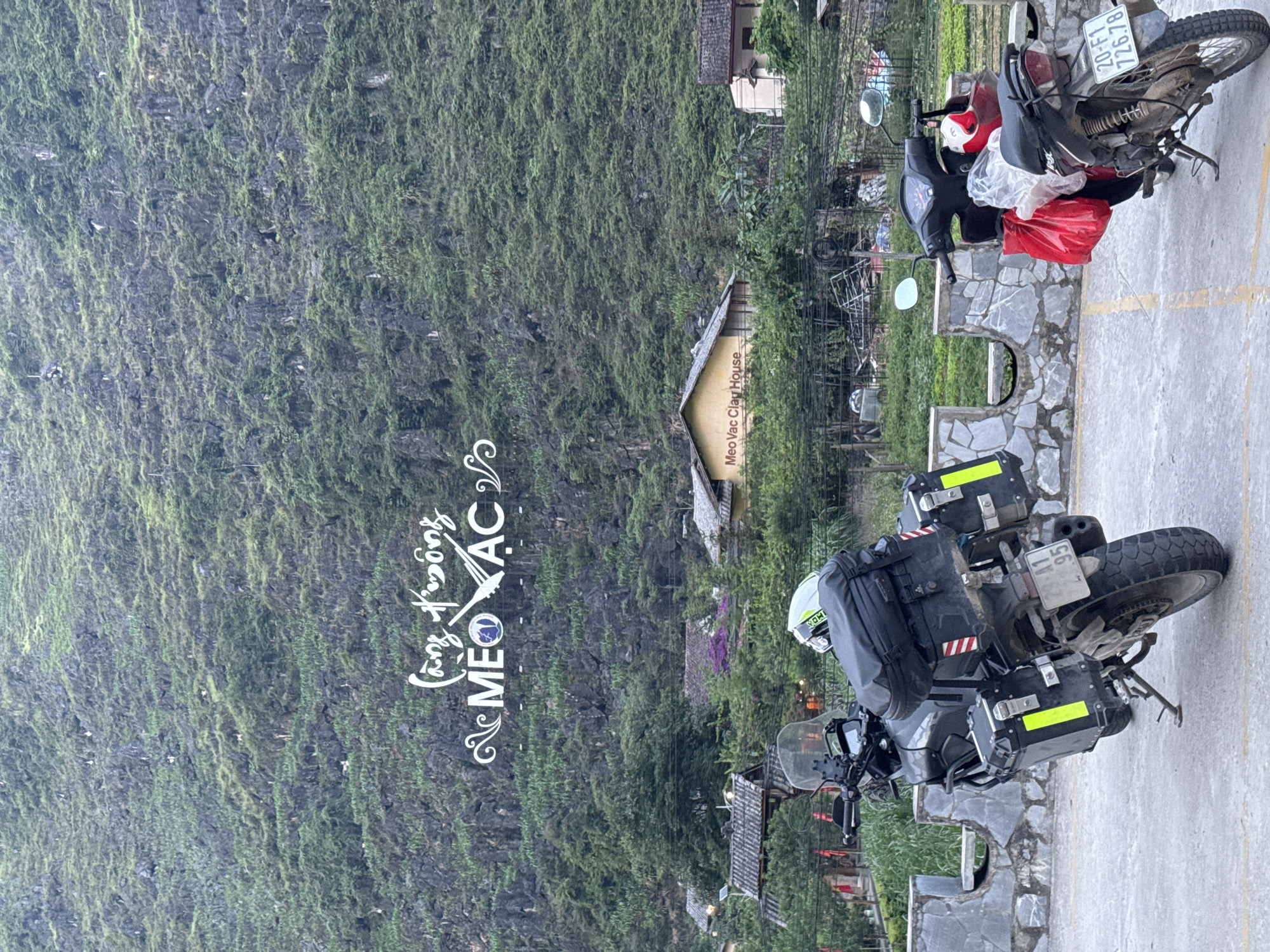 This photograph, taken in Mèo Vạc Commune, Vietnam, presents a landscape featuring a large, verdant, moss-covered mountain or cliff dominating the left side of the frame. The foreground showcases a partially paved area where two motorcycles are parked. One motorcycle is predominantly black, with storage boxes, a white helmet, and a license plate visible. The other motorcycle is a lighter color, possibly silver or gray, and appears more worn, with a red bag and a Vietnamese license plate number "20-F1 726.78" partially visible.
In the midground, there are structures and greenery indicating a settlement or developed area. A sign reads "Meo Vac Clay House". The overall setting implies a mountainous region in Vietnam, with a blend of natural terrain and human development.